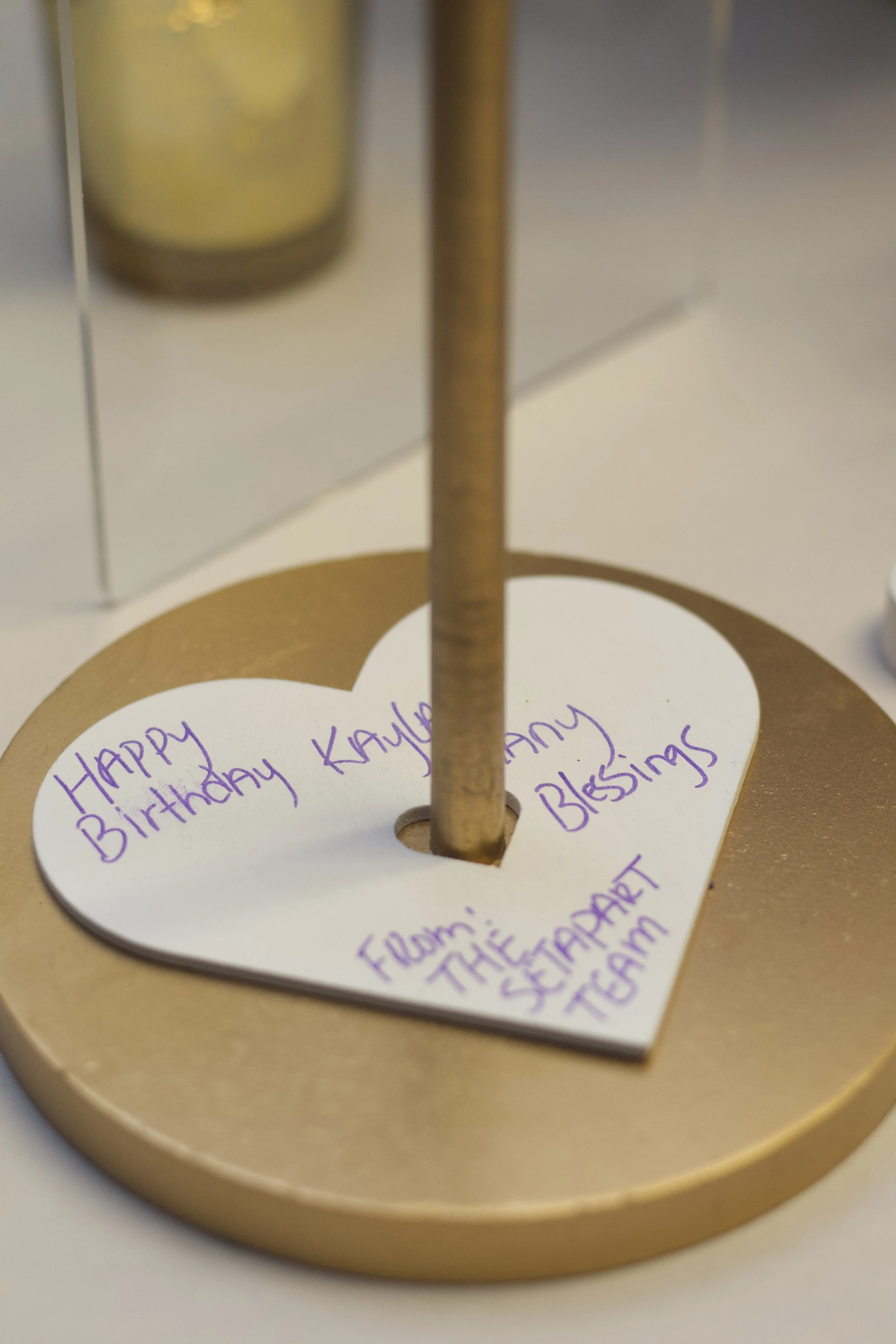 A gold-colored heart-shaped note with a handwritten message in purple ink, reading: 'Happy Birthday Kayla. Many blessings! From: the sermon team.' The note is attached to a gold circular base with a central hole, supporting a vertical rod.