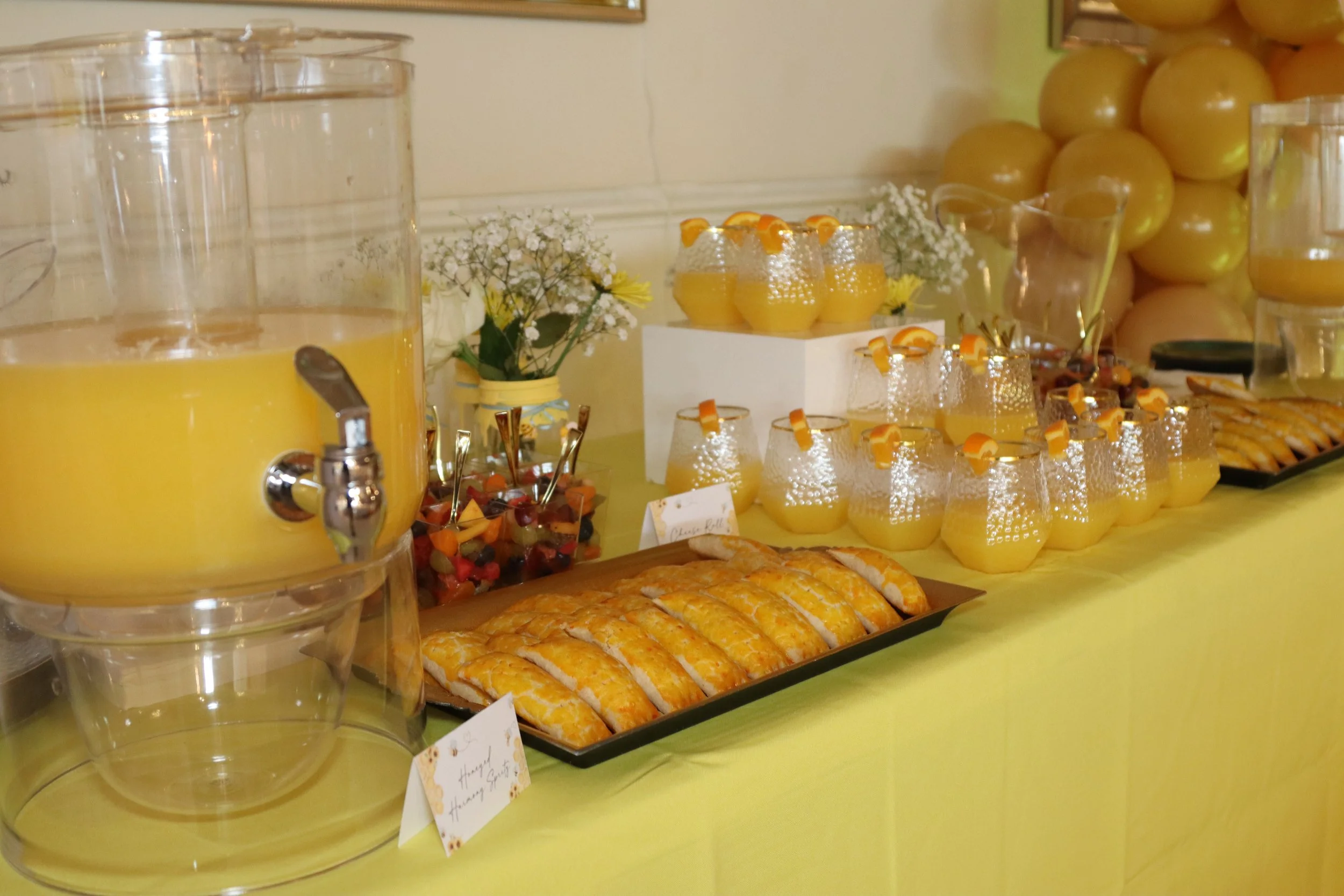 A table set up with yellow drinks in large dispensers, glasses of yellow juice topped with orange peel, a tray of glazed pastries, fruit skewers, and decorative flowers in a yellow vase, with gold balloons in the background.