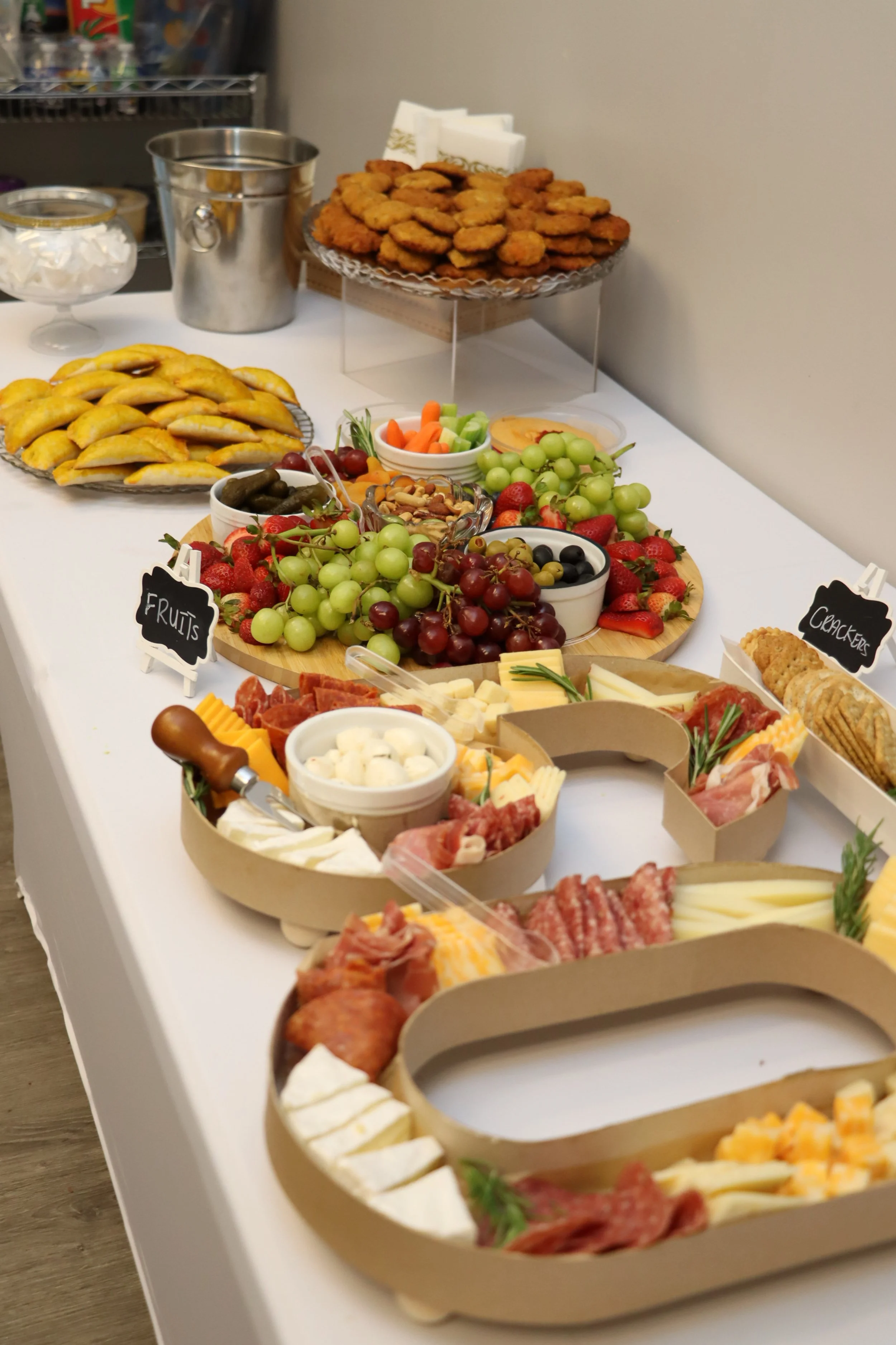 A cheese and charcuterie board with grapes, strawberries, and nuts, along with a platter of fruit, crackers, and breadsticks, set on a white table. In the background are fried chicken, mini sandwiches, and a variety of dipping sauces.