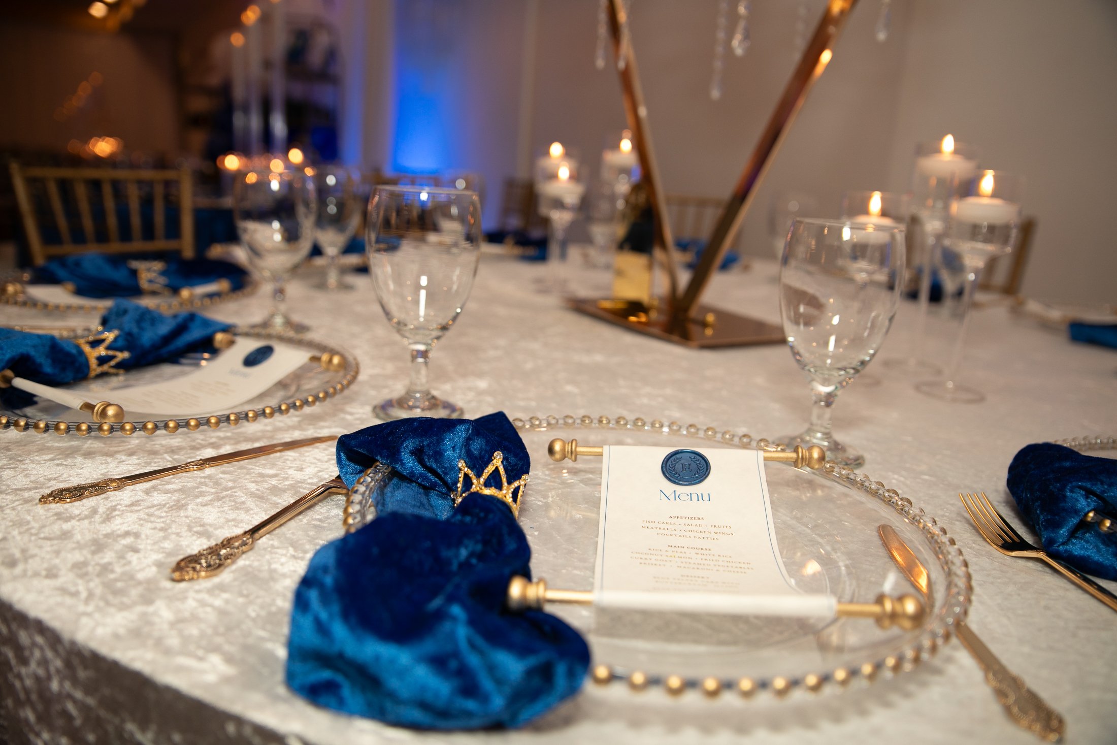 Elegant banquet table set with blue napkins, gold beaded chargers, clear glassware, and candles in the background.