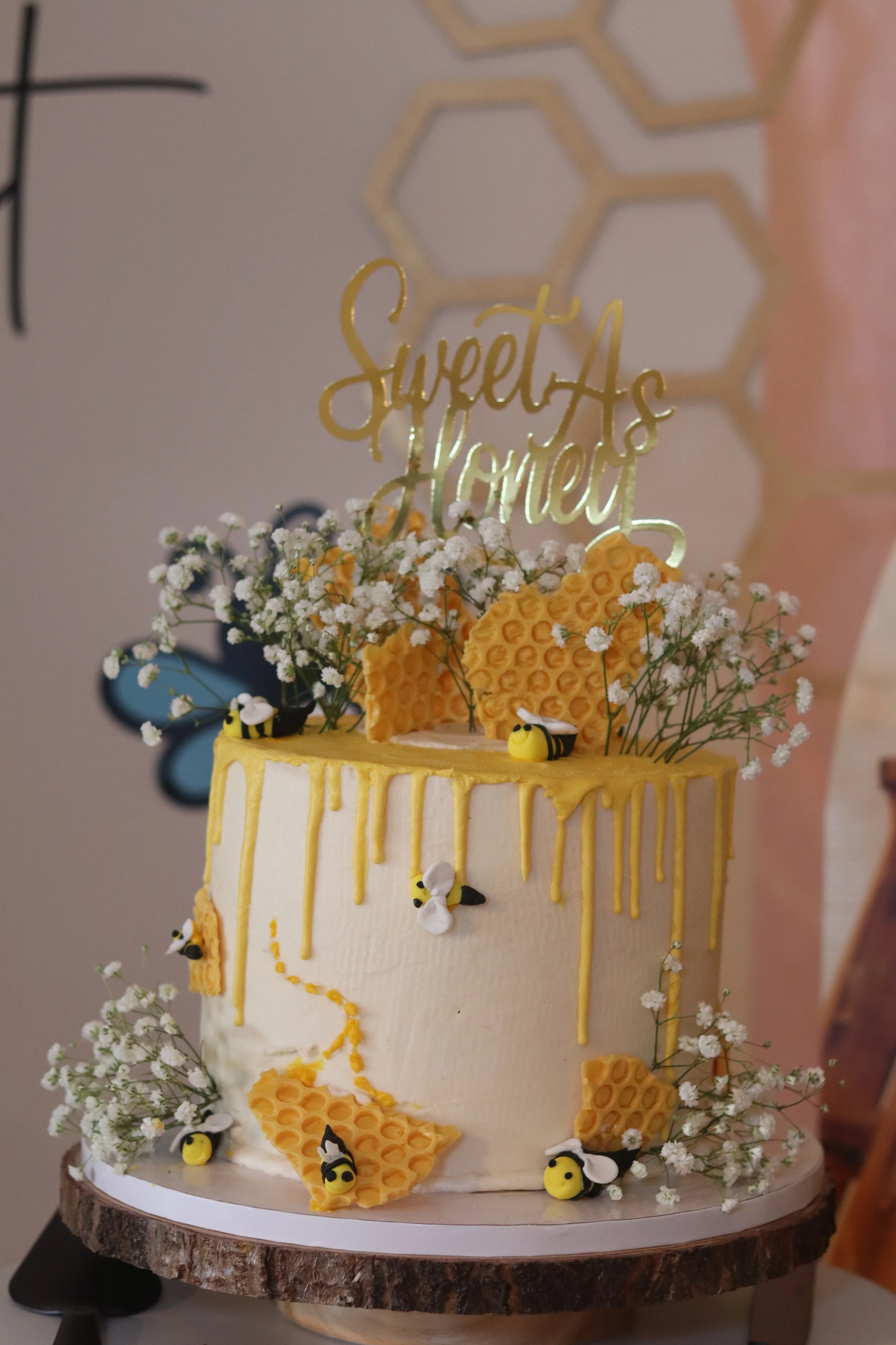 A honey bee themed cake with white frosting, honeycomb decorations, small bees, white flowers, and a gold cake topper that says 'Sweet As Honey.'