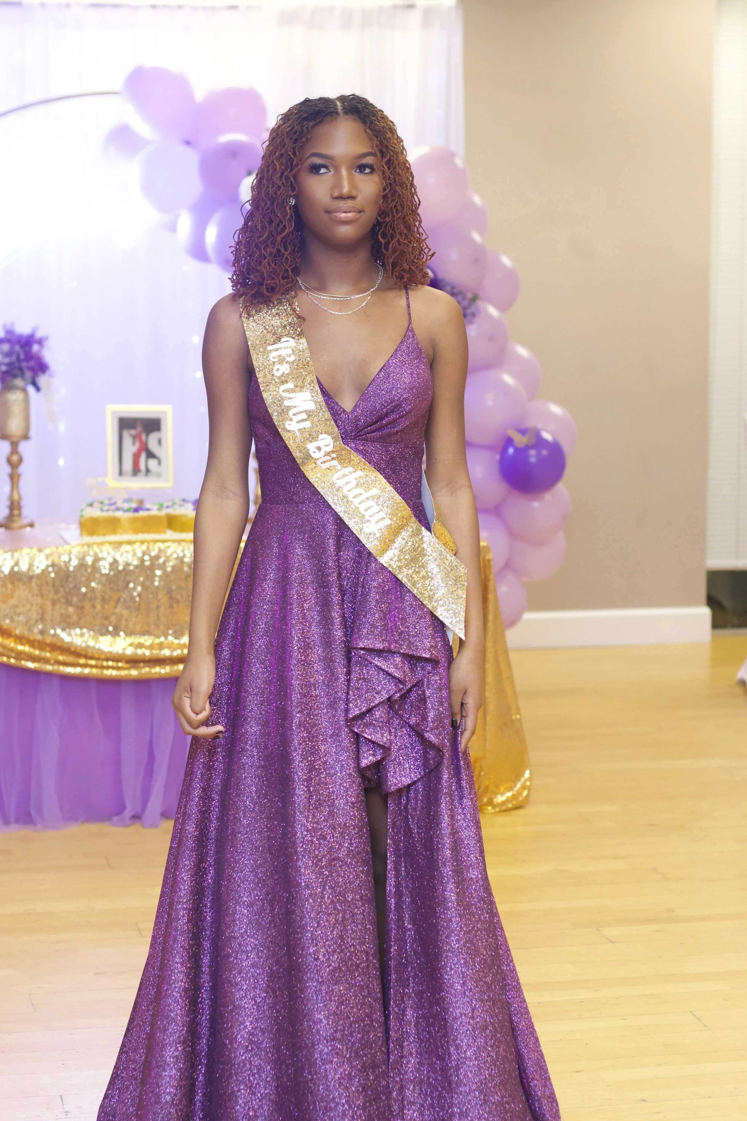 Young woman in a purple, glittery dress with a sash that reads '18th My Birthday' standing at a celebration with purple and pink balloons and a decorated table in the background.