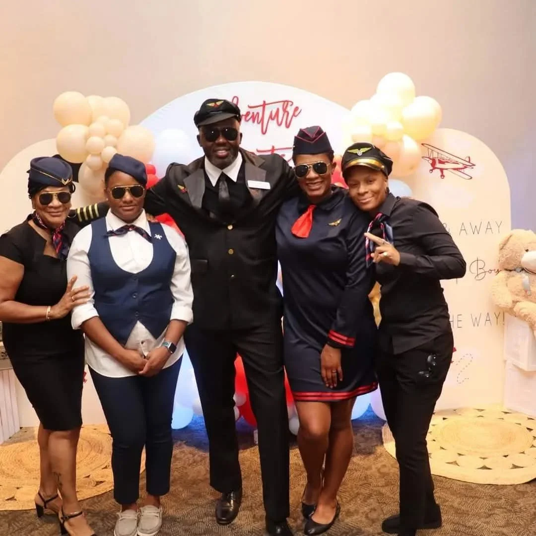 Group of five people dressed as airline crew members standing together, some wearing pilot hats and aviator sunglasses, with a backdrop featuring balloons and an airplane illustration.