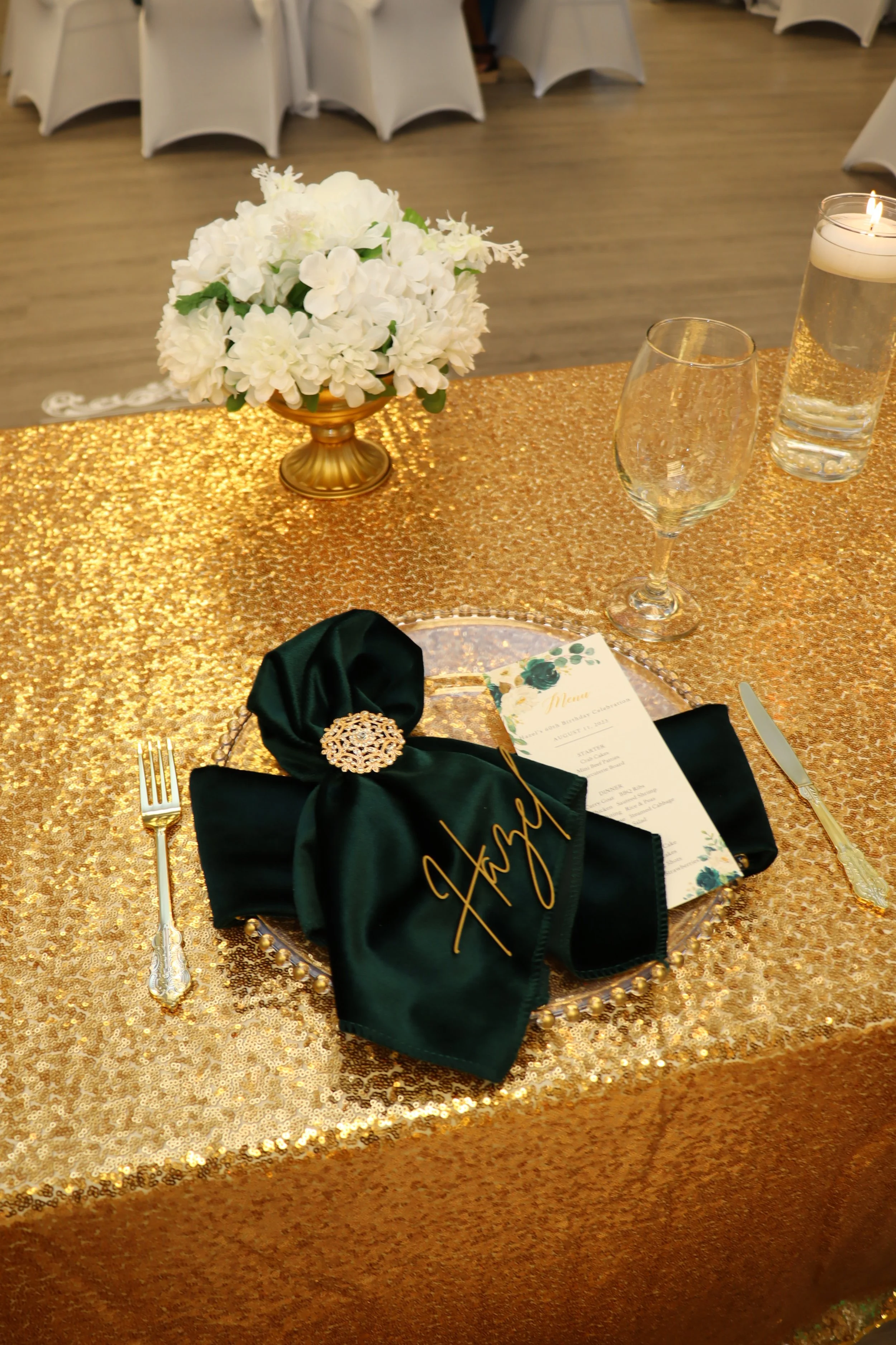 A decorated event table with a gold sequin tablecloth, a dark green napkin with gold embroidery and a decorative brooch, a clear flower plate with a menu, a gold fork and knife, a flower centerpiece in a gold vase, and two glasses, one with wine and 