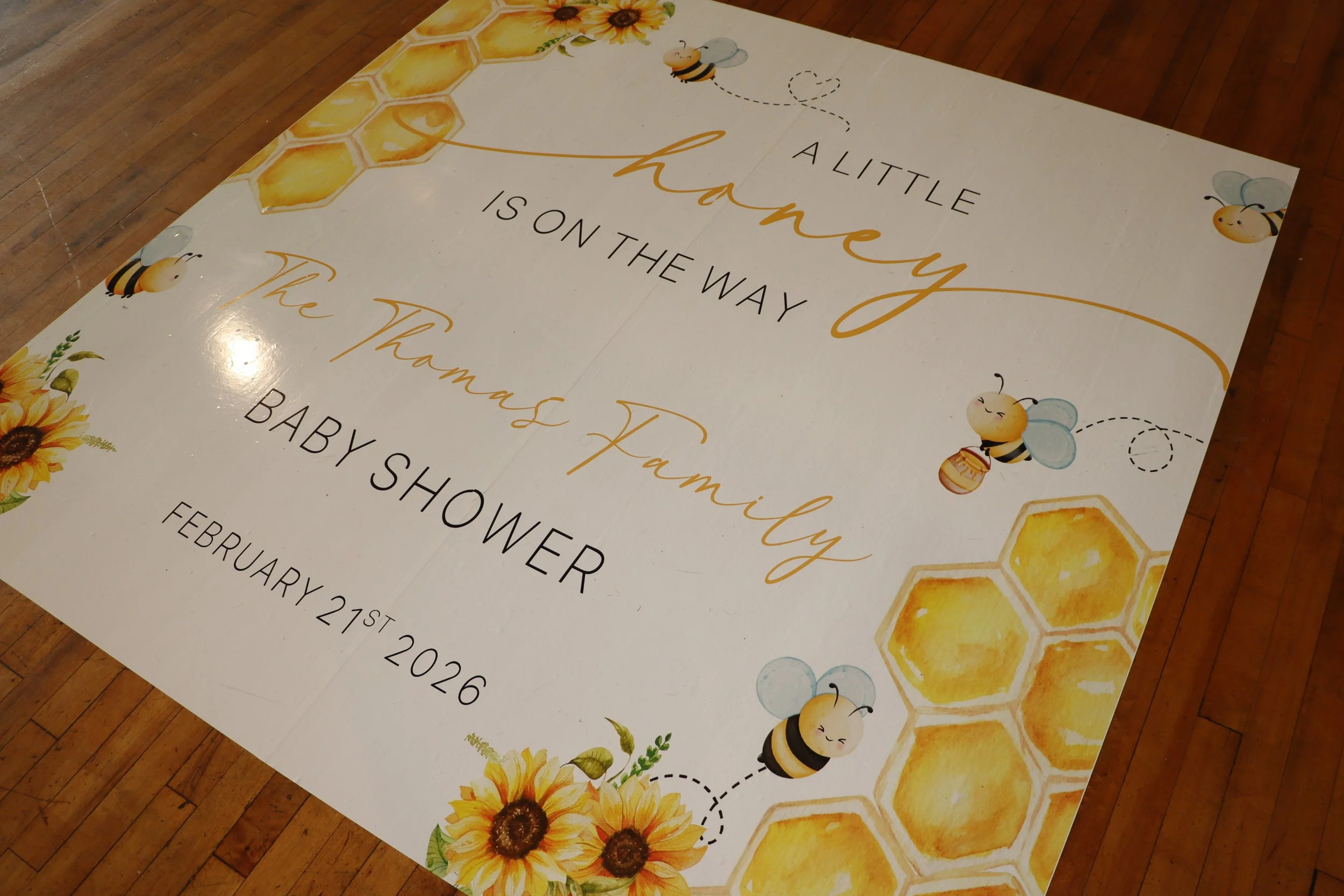 Decorative poster for a baby shower with honeybee and honeycomb illustrations, flowers, and text that reads 'A little honey is on the way. The Thomas Family. Baby Shower. February 21st, 2026.'