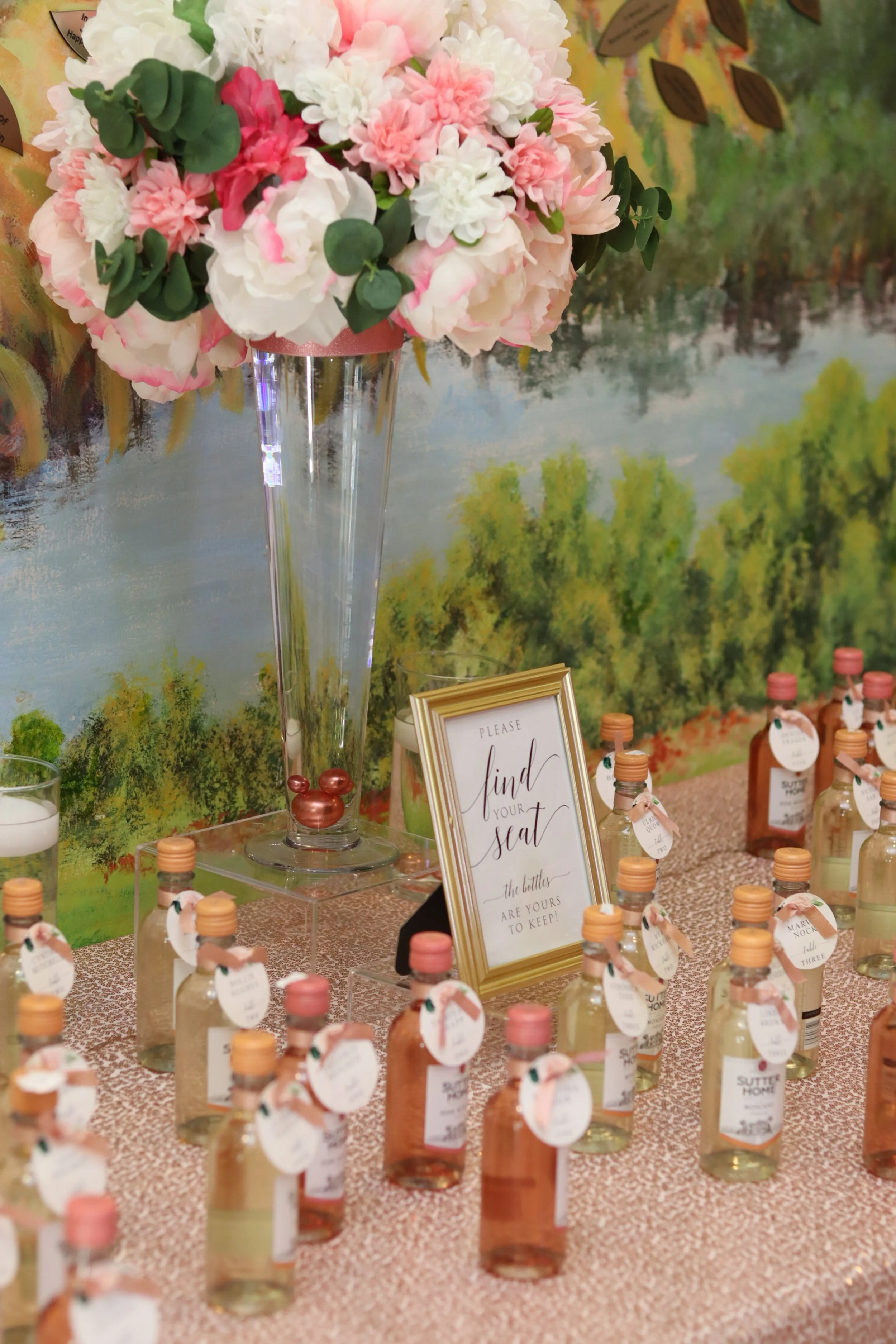 A large bouquet of pink and white flowers with greenery in a tall glass vase on a decorated table with small bottles of bubble solution, and a sign that reads 'Please find your seat, the bottles are yours to keep.'