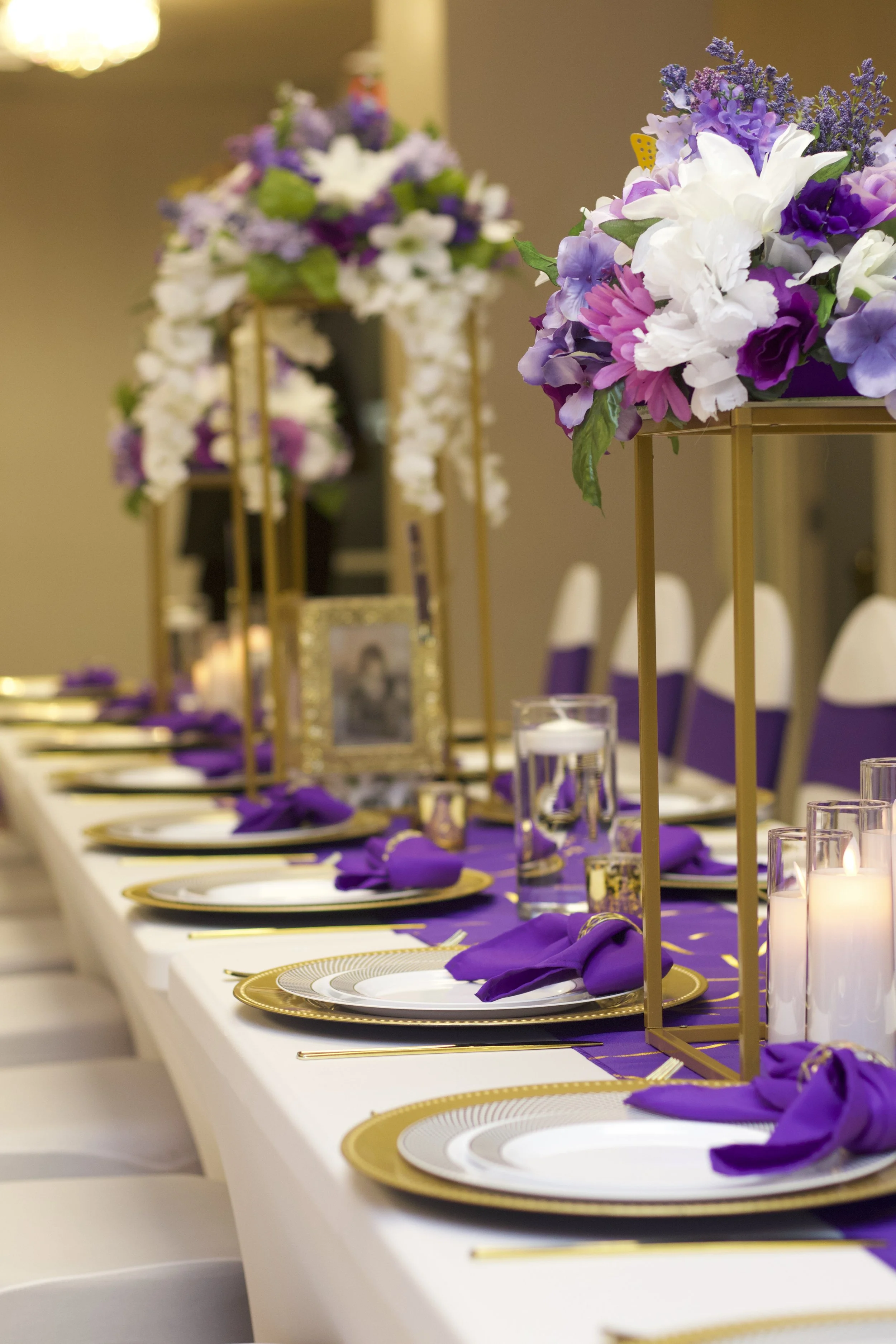 Elegant banquet table decorated with purple and white floral centerpieces, gold-rimmed plates, purple napkins, candles, and glassware.