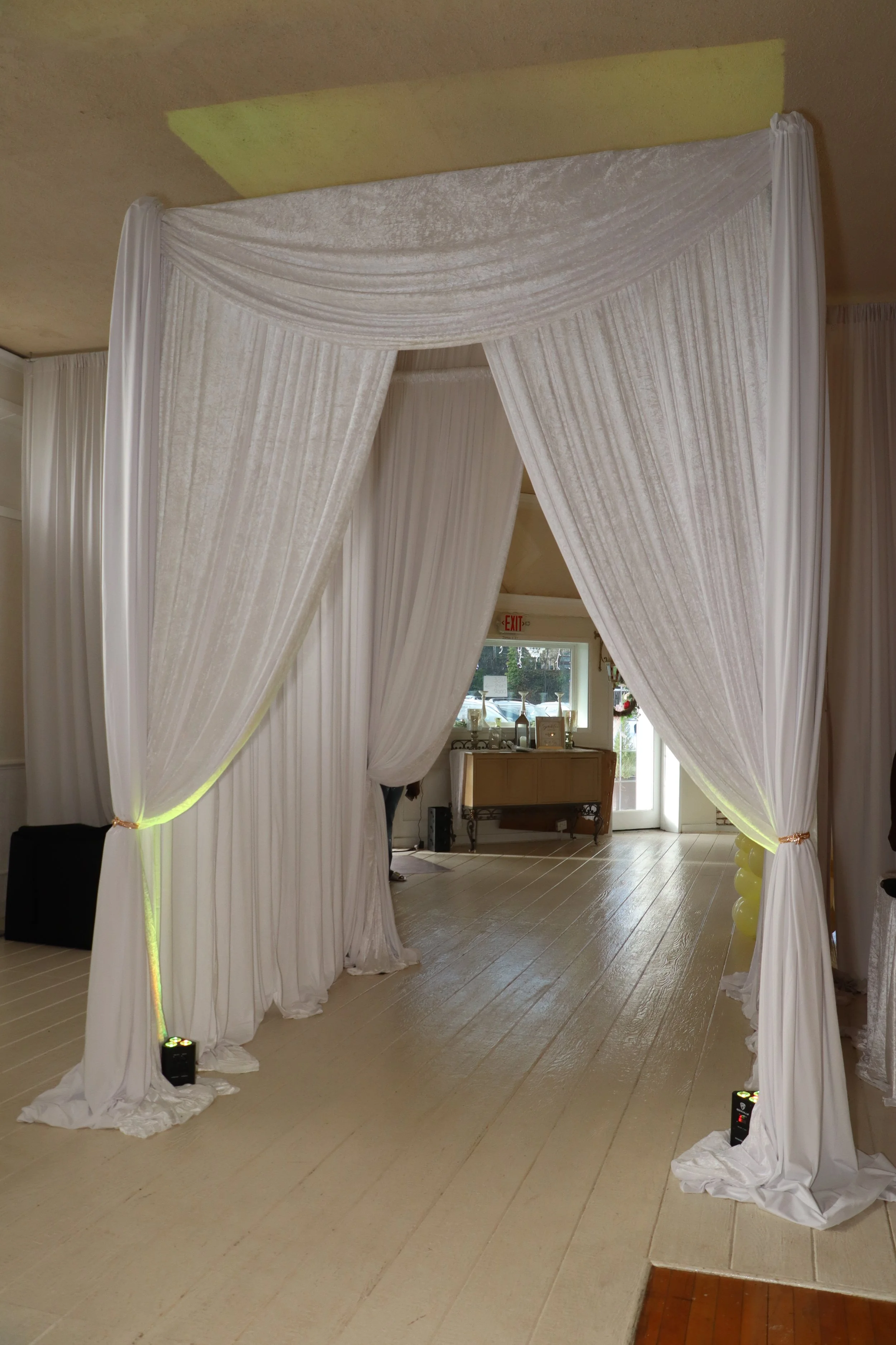 White draped fabric archway or tent, set up indoors for a wedding or event, with lighting at the base of each side.