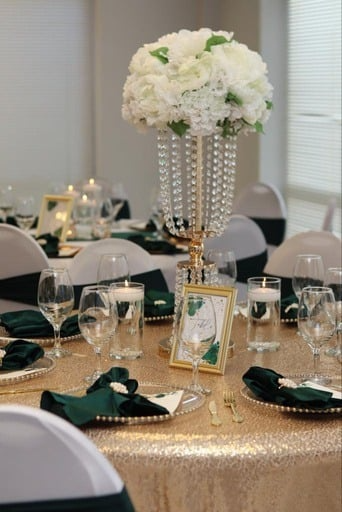 Table decorated with a tall floral centerpiece of white flowers in a jeweled vase, surrounded by elegant place settings with black napkins, glassware, and gold flatware, in a bright room with window blinds.