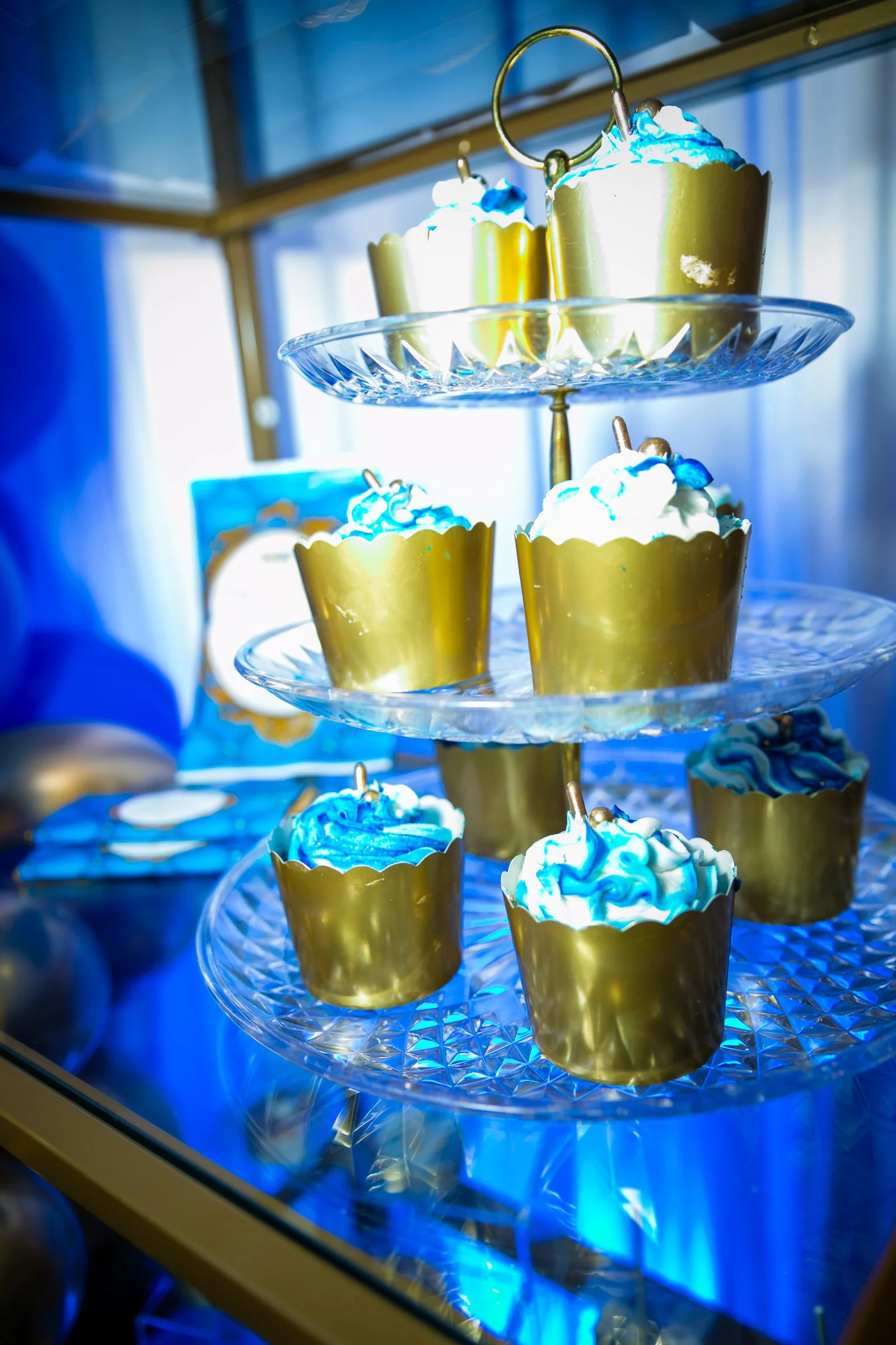 Cupcakes with white and blue frosting on a glass display stand with blue lighting.