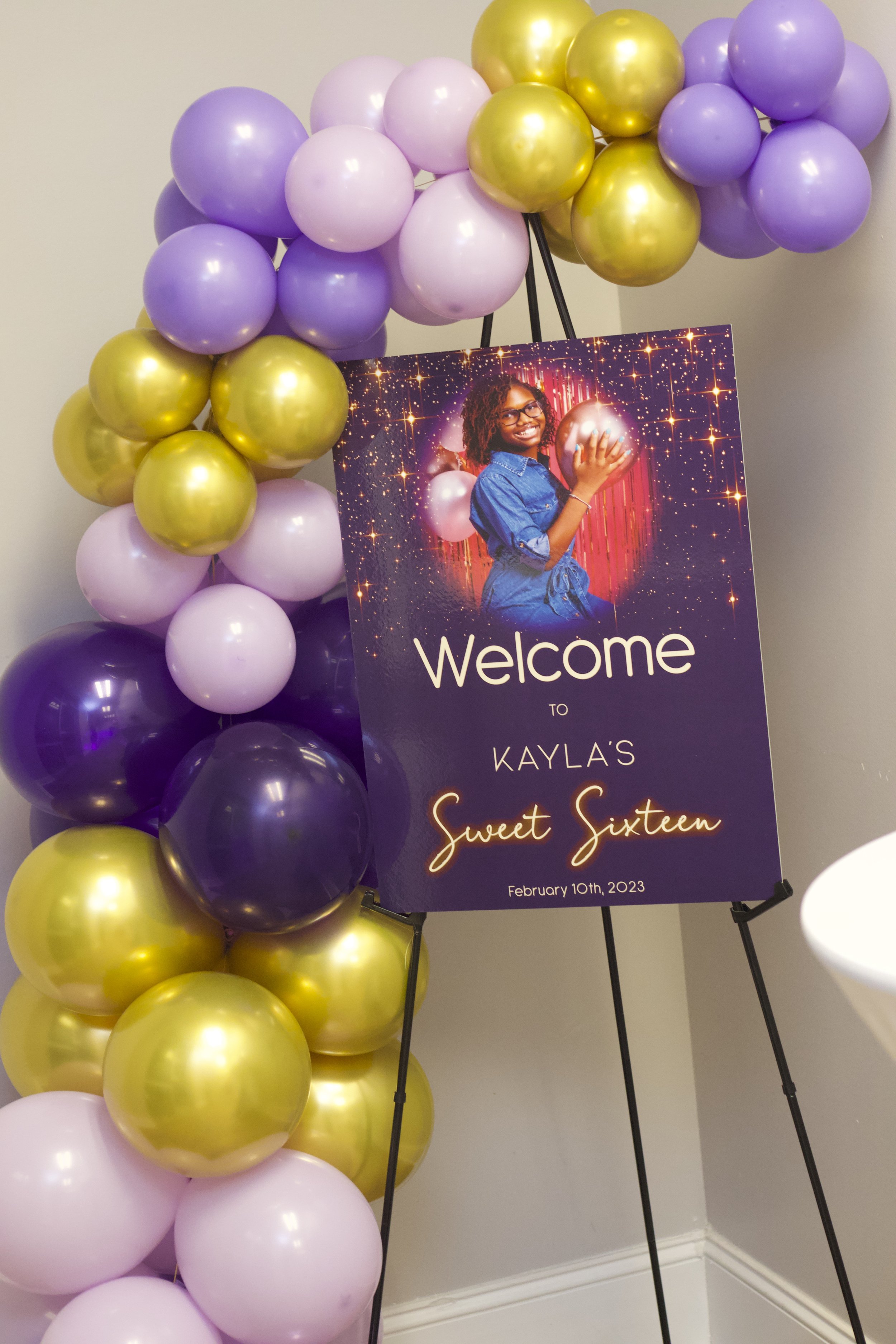 Celebration welcome sign for Kayla's 16th birthday, featuring a girl holding a shiny balloon, surrounded by purple and gold balloons, with the date February 10th, 2023.