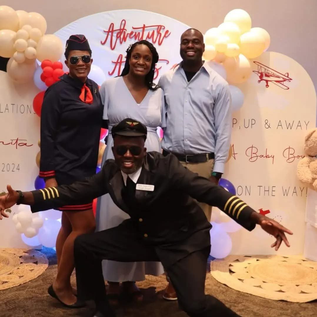Four people posing together at a themed event with a backdrop that says "Adventure Awaits." Two men and two women, with one man dressed as a pilot and one woman dressed as a flight attendant. The backdrop is decorated with balloons and an airplane gr
