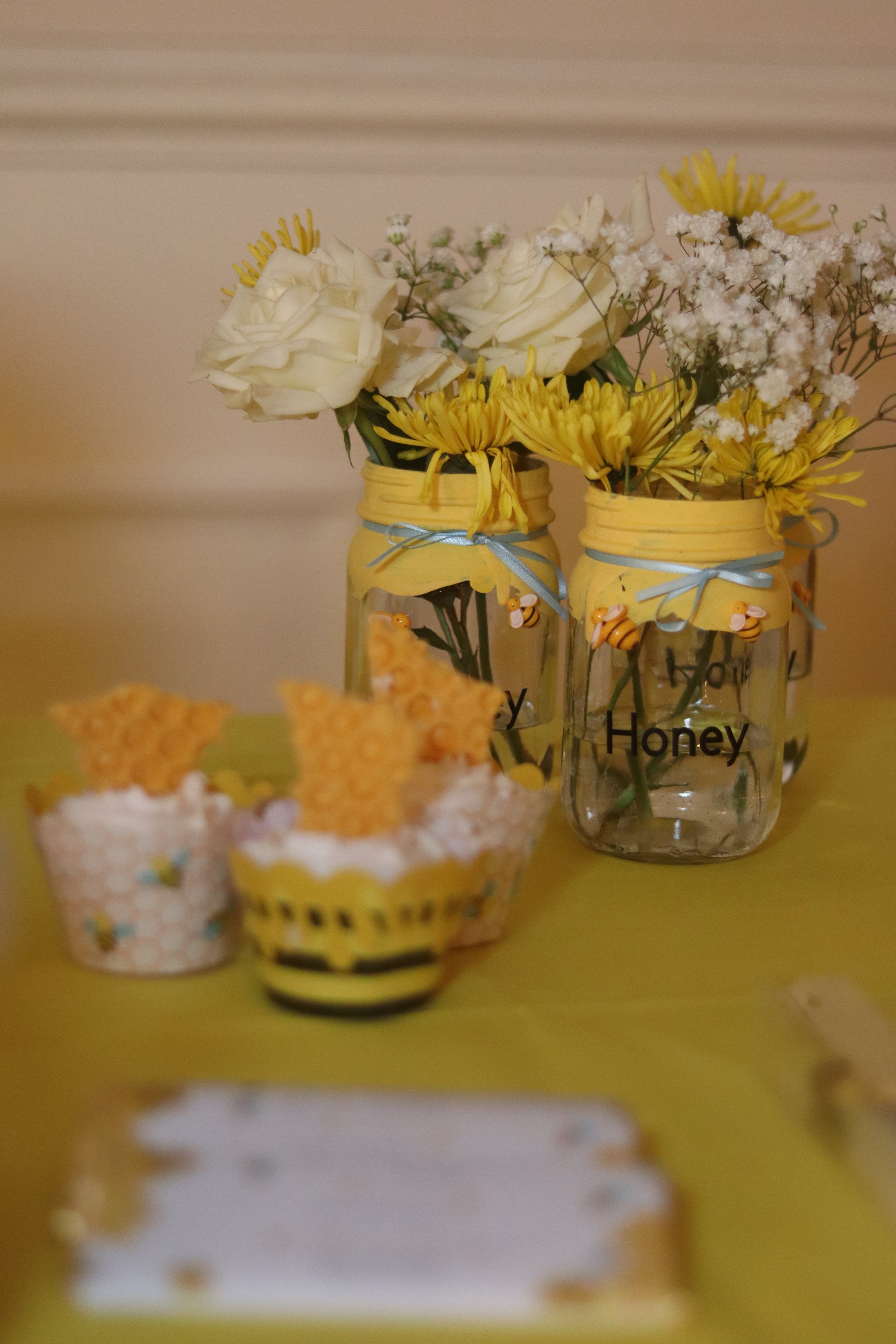 Decorative jars with yellow fabric covers and ribbons filled with white roses, yellow daisies, and white baby's breath on a yellow table with honey-themed cupcake decorations.