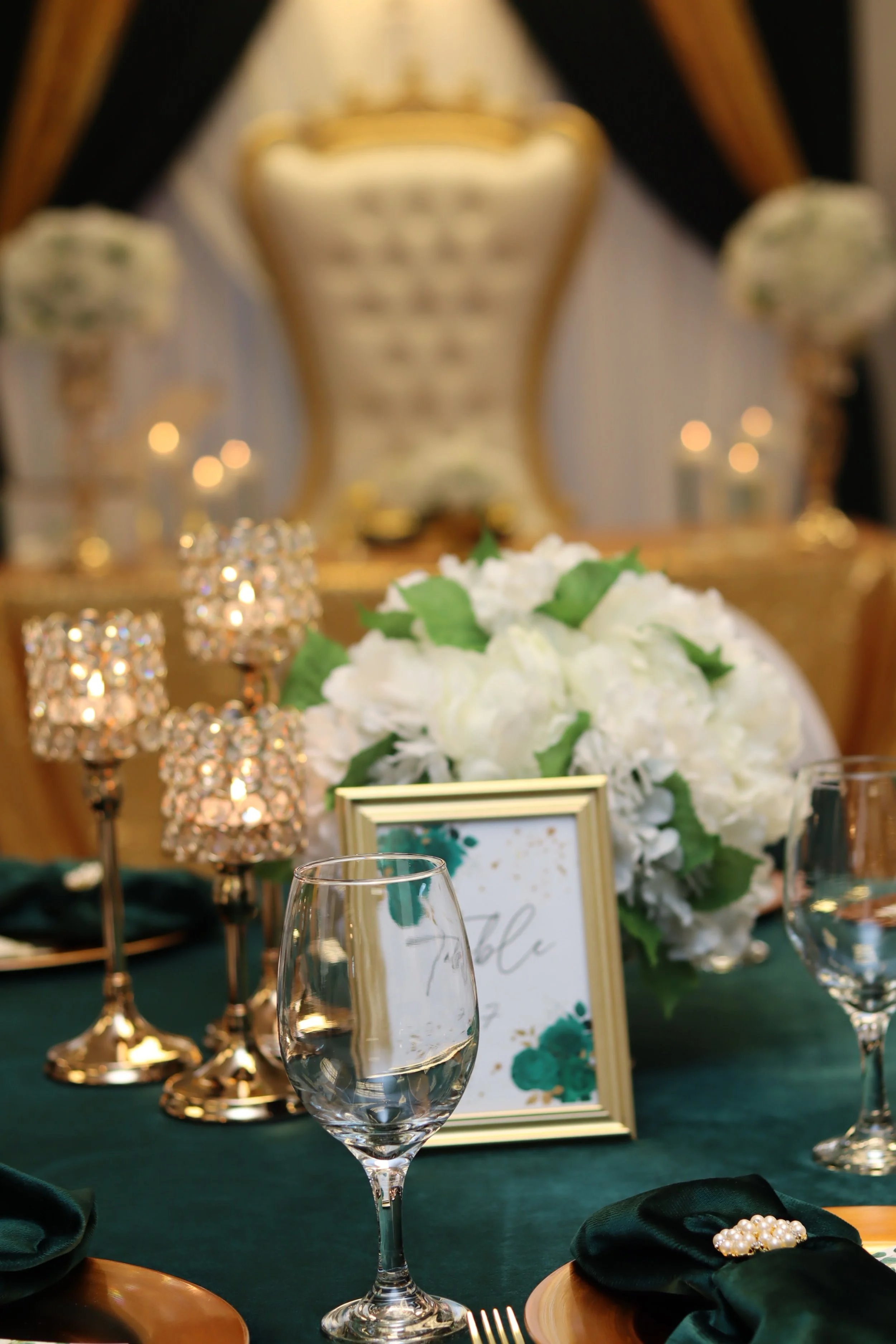 Elegant wedding reception table with white floral centerpiece, gold framed sign, crystal candle holders, empty glasses, and green and gold accents.