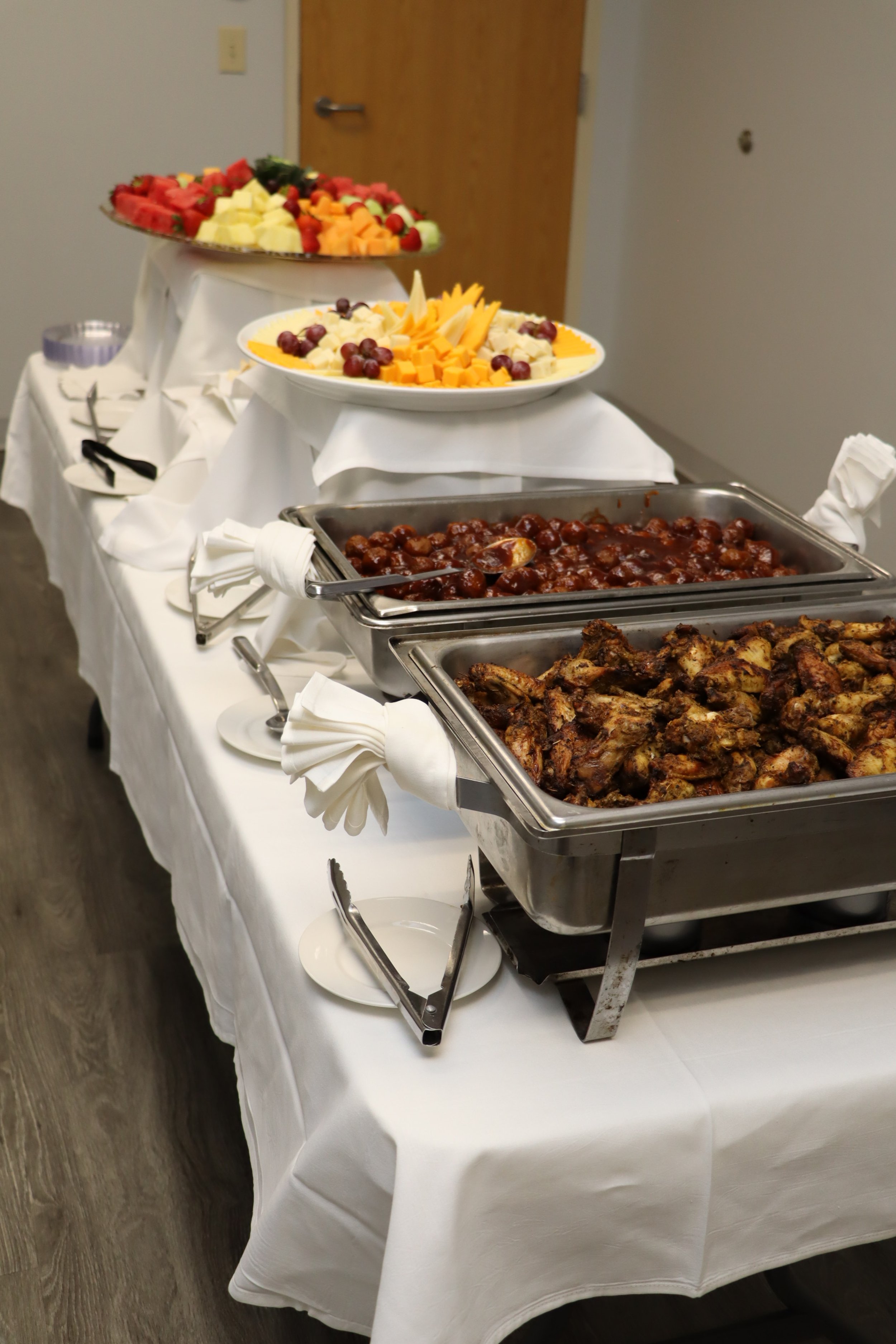 Buffet table with trays of chicken, meatballs in sauce, and sliced cheese and fruits, along with bowls of fresh fruit and a plate of grapes.