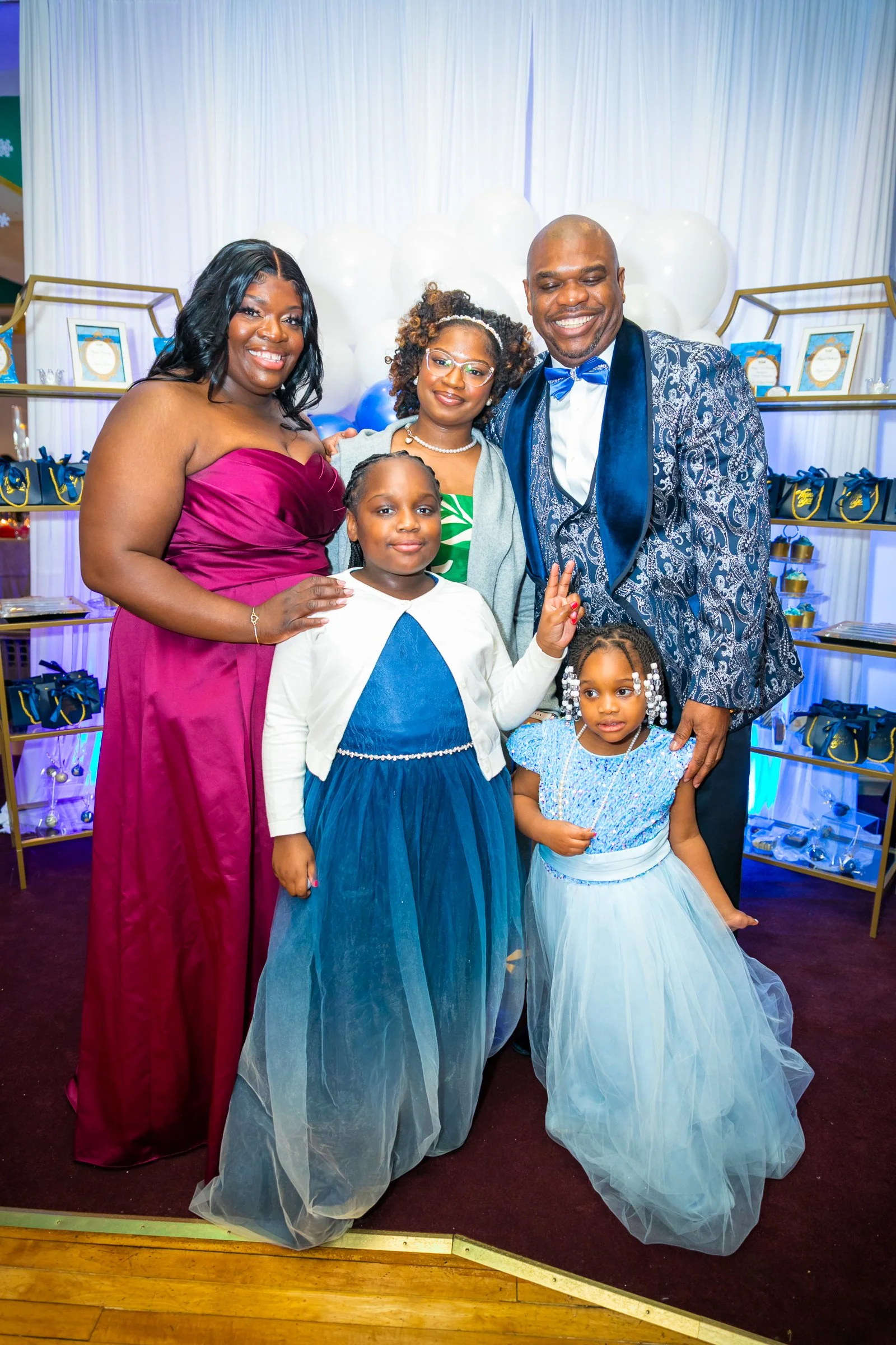 Group of six people, four women and two young girls, smiling and posing at a celebration event with decorations and white balloons in the background.