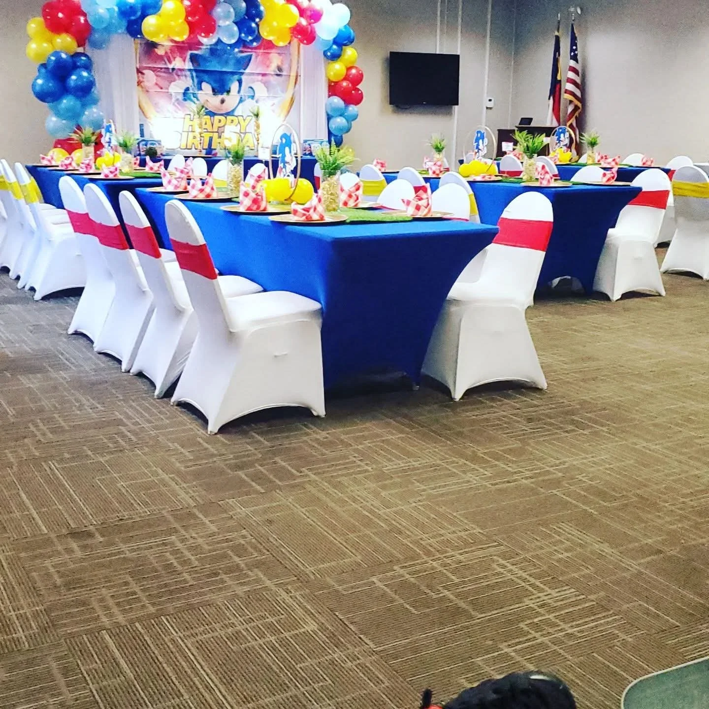 Decorated banquet hall with a blue, red, and yellow theme for a birthday party, featuring a table with themed tableware, pineapple centerpieces, and balloon arrangements with a Sonic the Hedgehog backdrop.