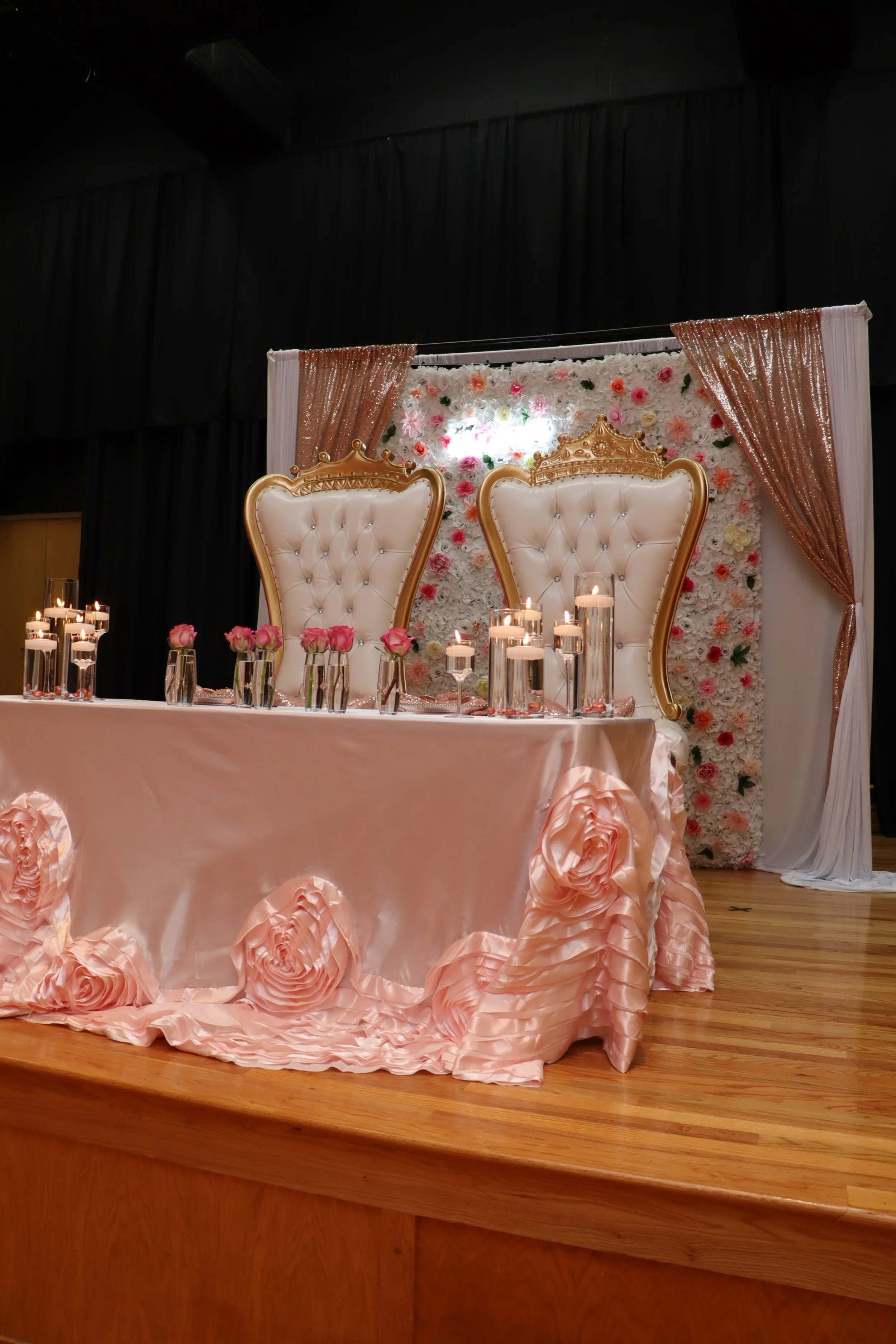 Elegant wedding or event reception table with two ornate high-backed chairs, surrounded by pink roses and candles, set against a floral backdrop with pink and white flowers, gold accents, and draped curtains.