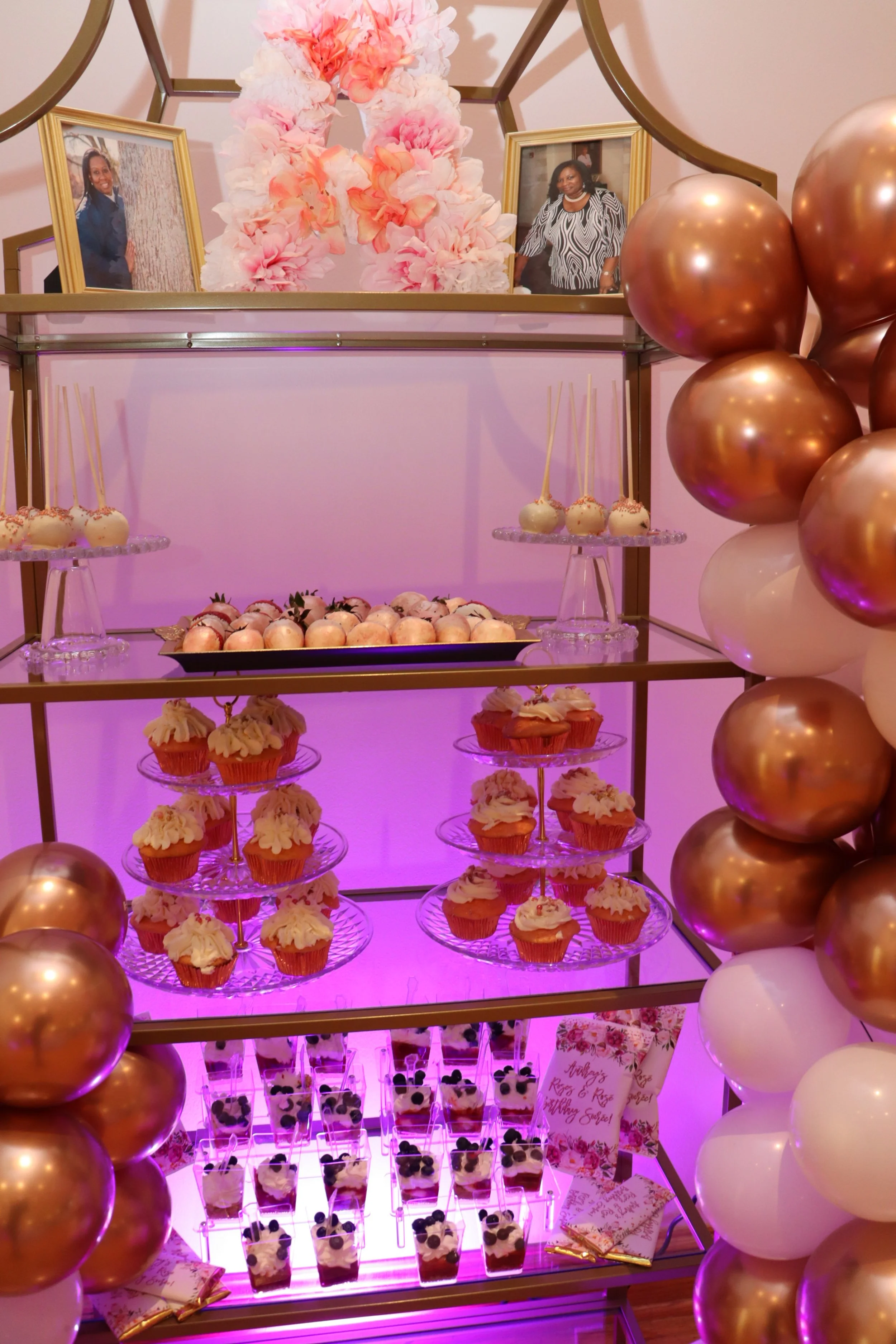 Dessert display with cupcakes, cake pops, and layered parfaits, decorated with pink and gold balloons and framed photos, at a celebration event.