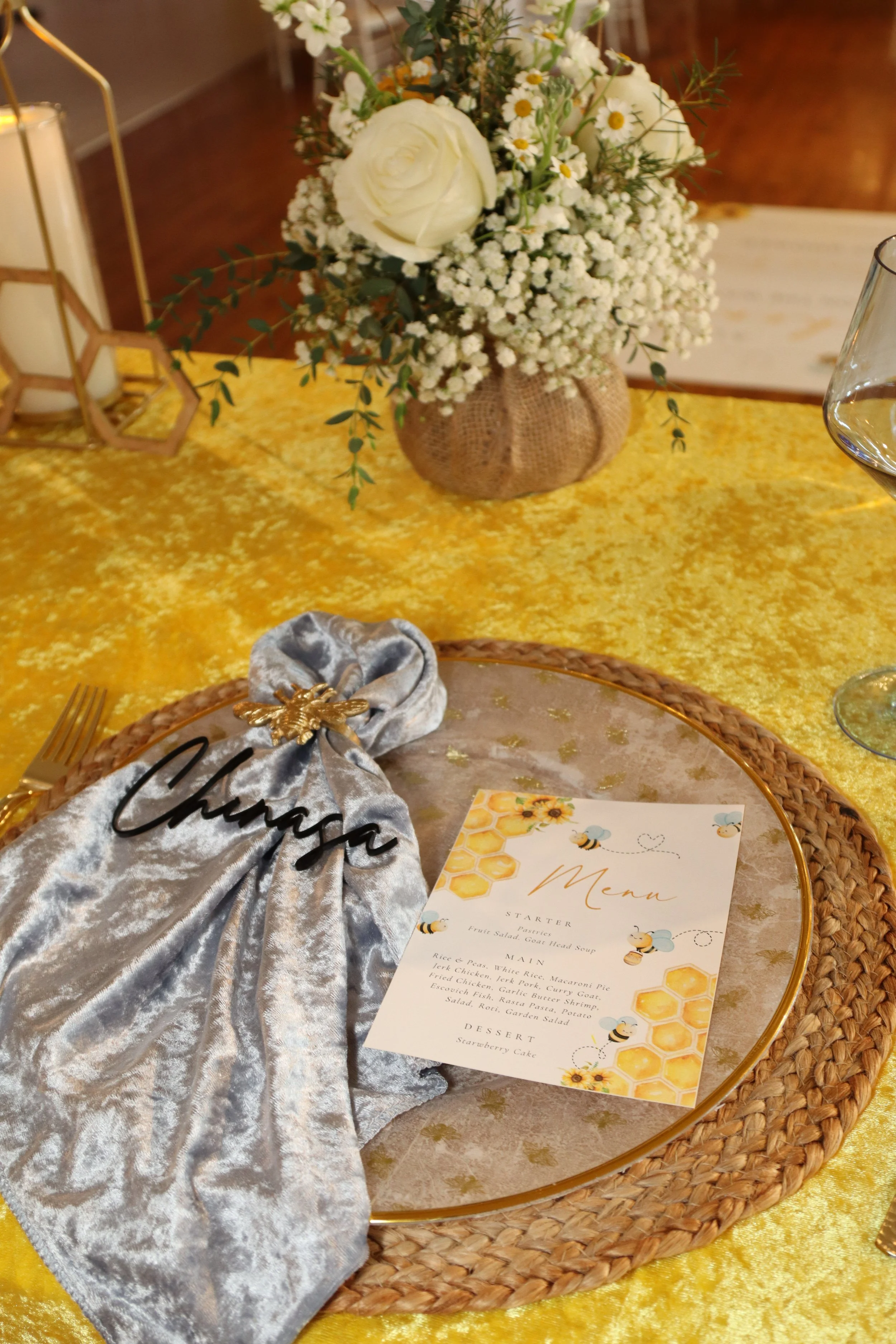 A table setting with a round glass plate, a menu card featuring bees and honeycomb design, a silver velvet cloth with a black name tag 'Chunga,' a floral arrangement with white roses and smaller white flowers in a burlap-wrapped vase, and a golden ta