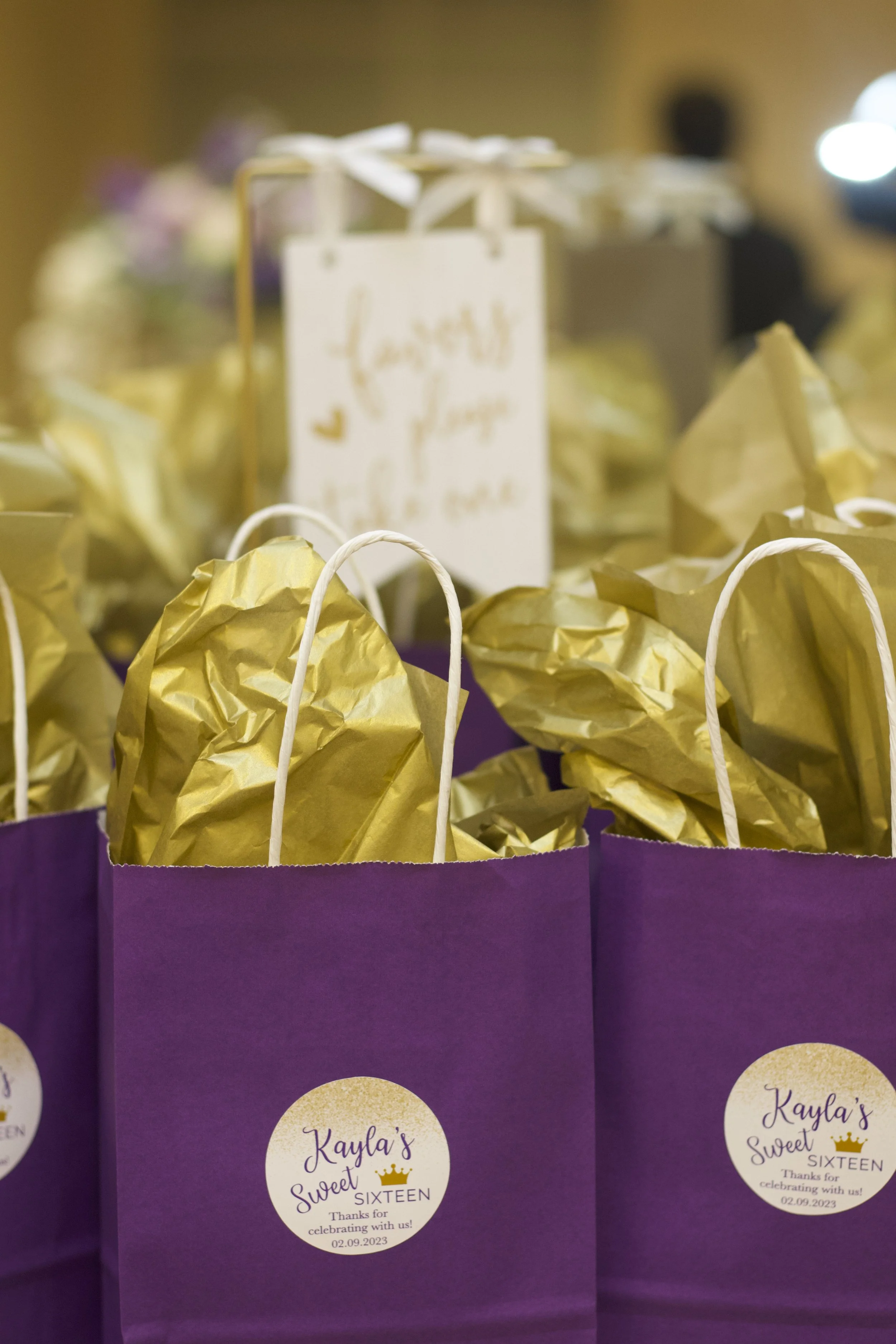 Purple gift bags with white handles and gold tissue paper, labeled 'Kayla's Sweet Sixteen' with a crown graphic, used for a birthday celebration.