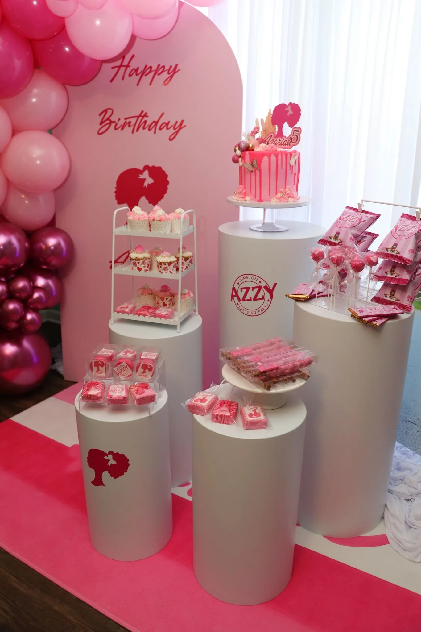 Pink-themed birthday party display with a cake, cupcakes, cookies, and party favors on white cylindrical pedestals. A backdrop reads 'Happy Birthday' with a silhouette of a girl, and balloons decorate the left side. Decorations are primarily in shade
