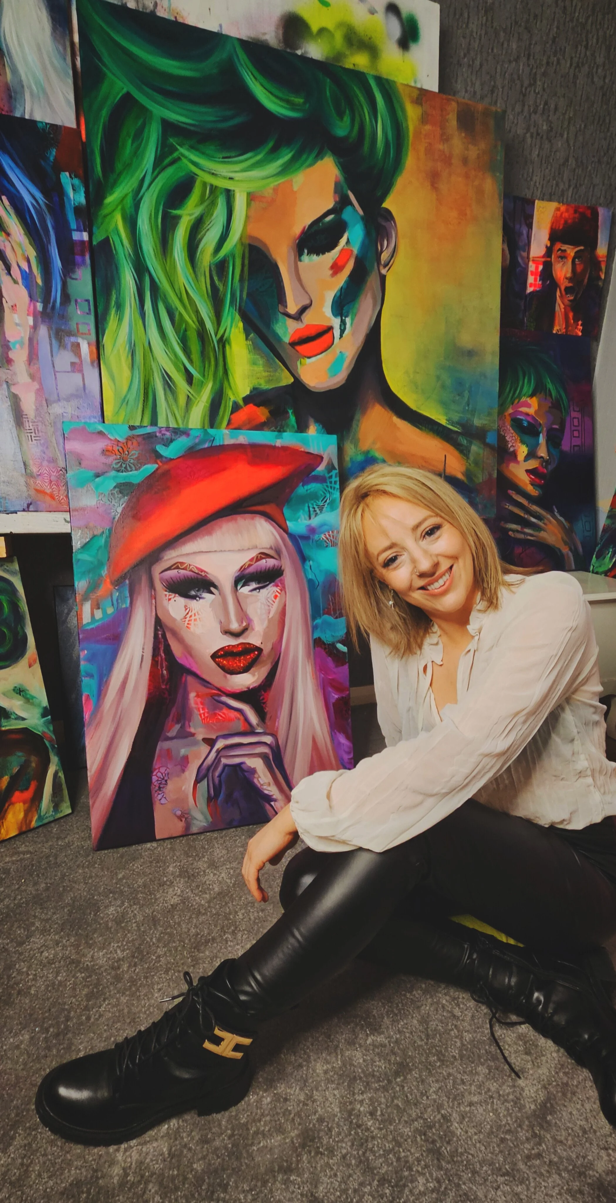 A smiling woman with blonde hair sitting on the floor in front of colorful, abstract portrait paintings of women, including one with green hair and others with vibrant makeup.