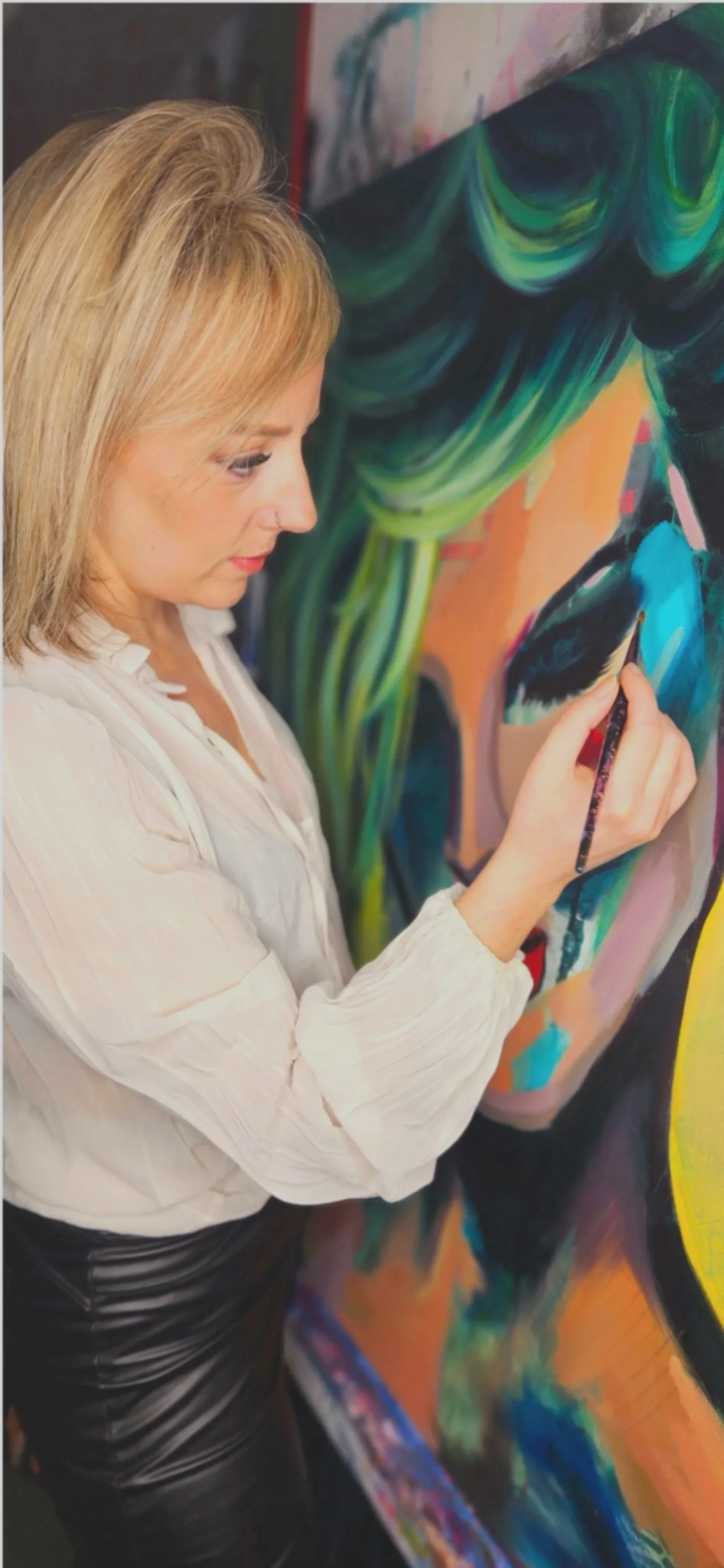 A woman with blonde hair, wearing a white blouse, painting a colorful abstract portrait on a large canvas.