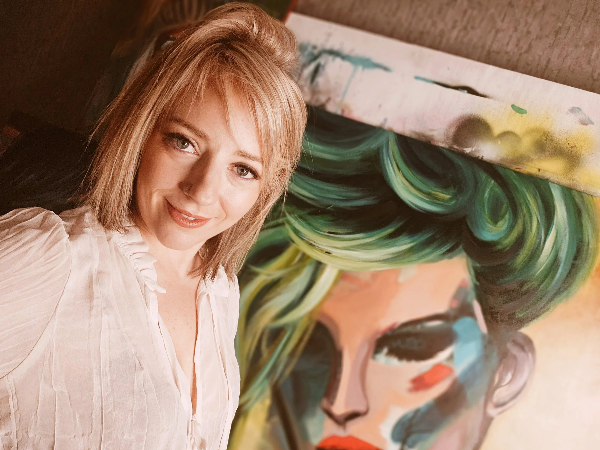 A woman with light skin, blonde hair, and wearing a light-colored blouse smiling at the camera, standing next to a colorful abstract painting of a face with green, black, and orange hair and bold makeup.