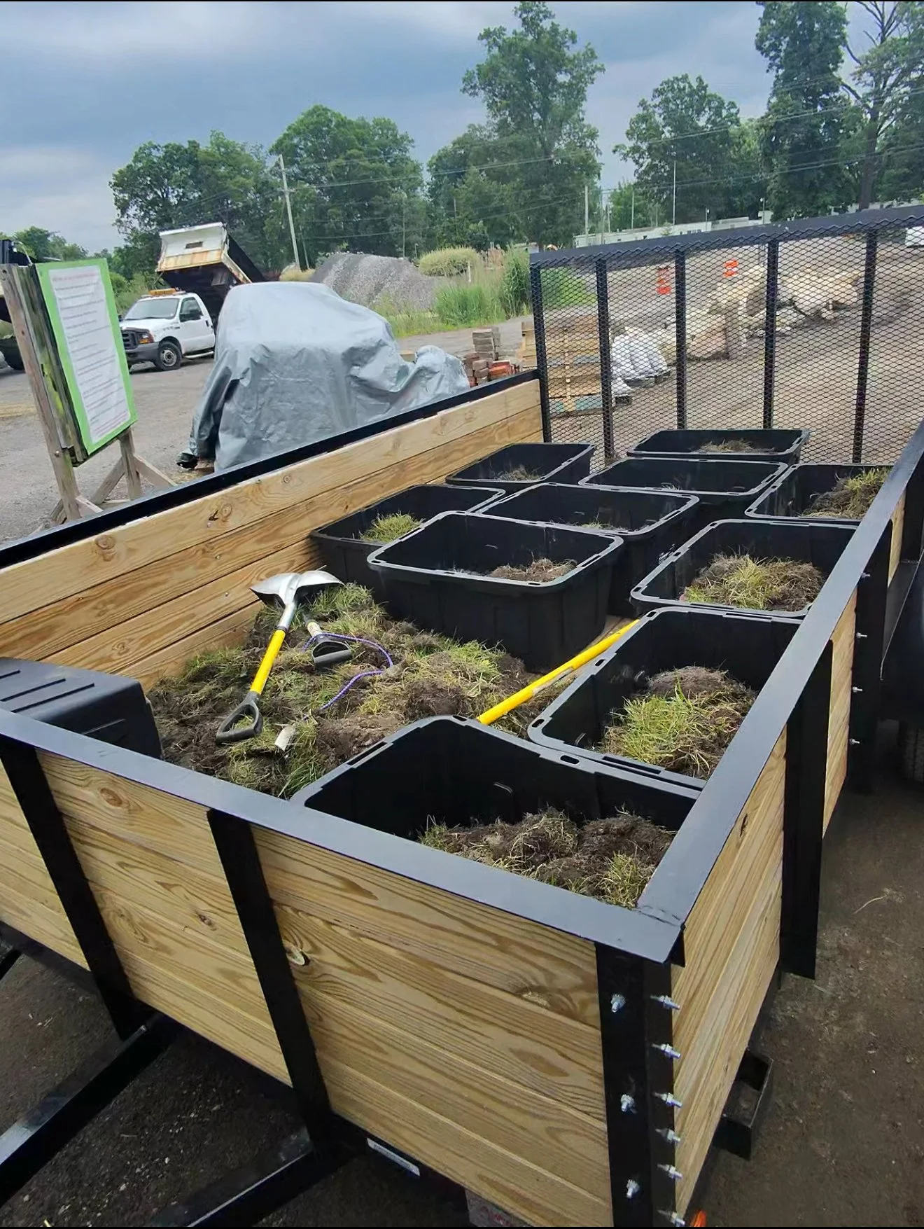A wooden raised garden bed filled with soil and young grass seedlings. Several black plastic containers with soil and grass are placed inside the bed. Gardening tools including a small spade with a yellow handle and a yellow rake are laid inside the bed. In the background, there are construction materials, parked vehicles, and trees under a cloudy sky.