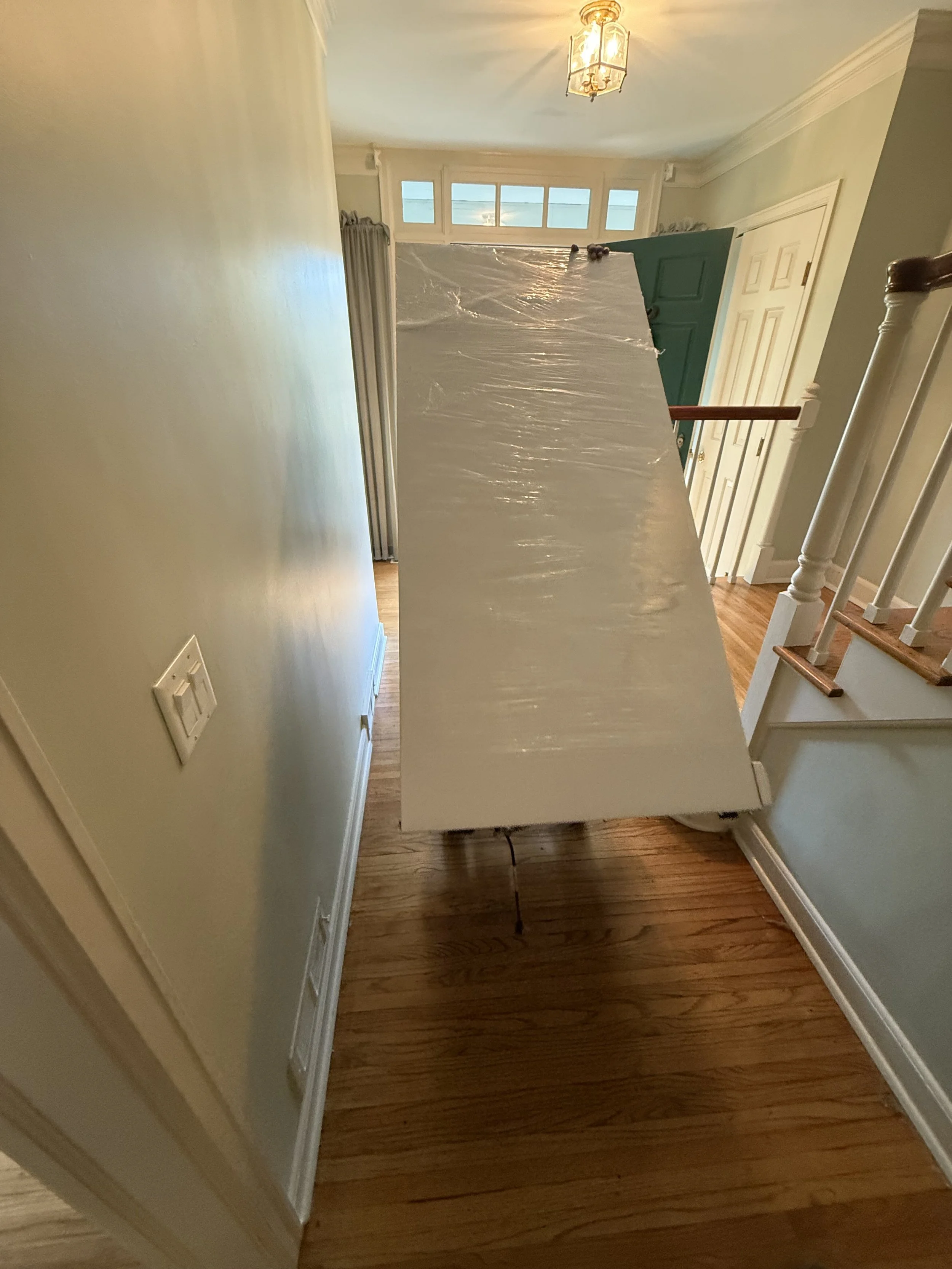 A large flat object covered with clear plastic wrap placed diagonally in a household hallway, with a staircase on the right and a door at the end of the hallway.