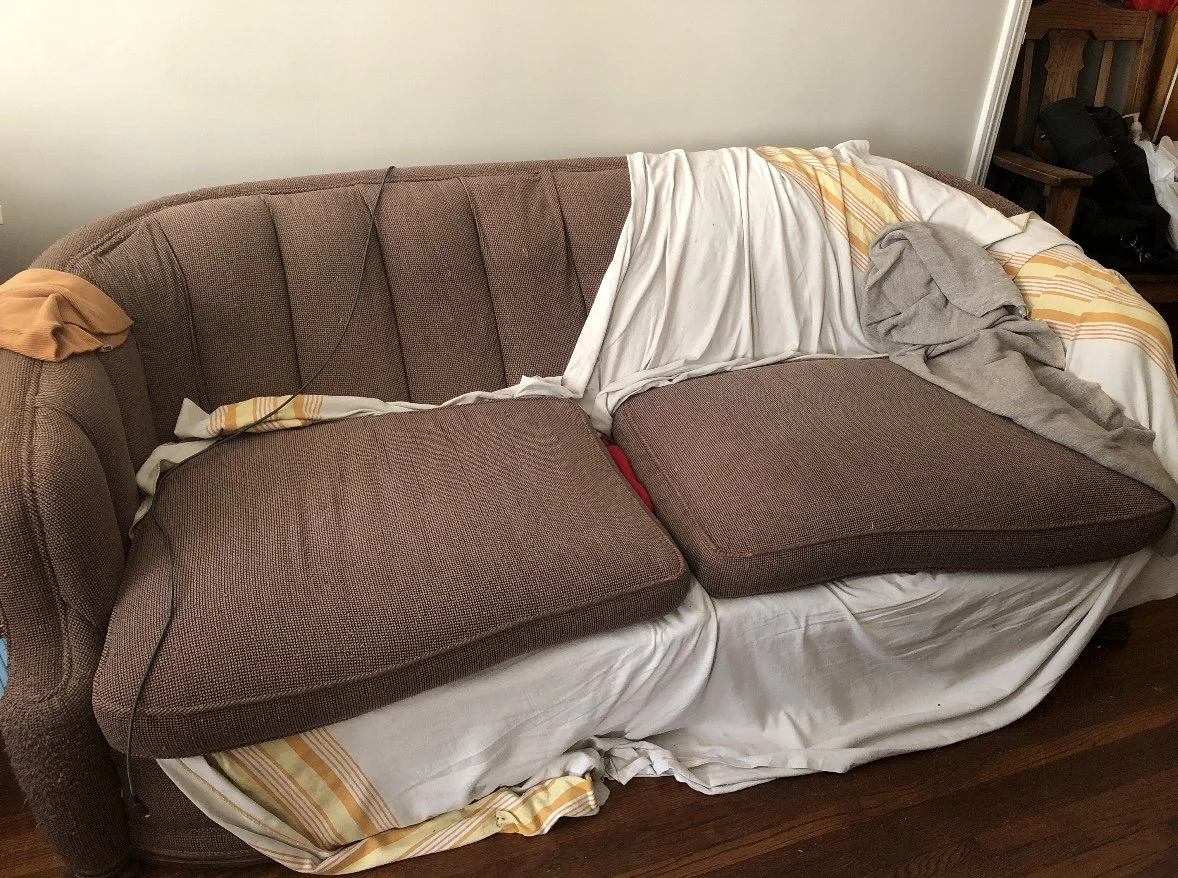 A brown upholstered sofa with two seat cushions, covered with bedsheets and clothing, positioned against a wall.