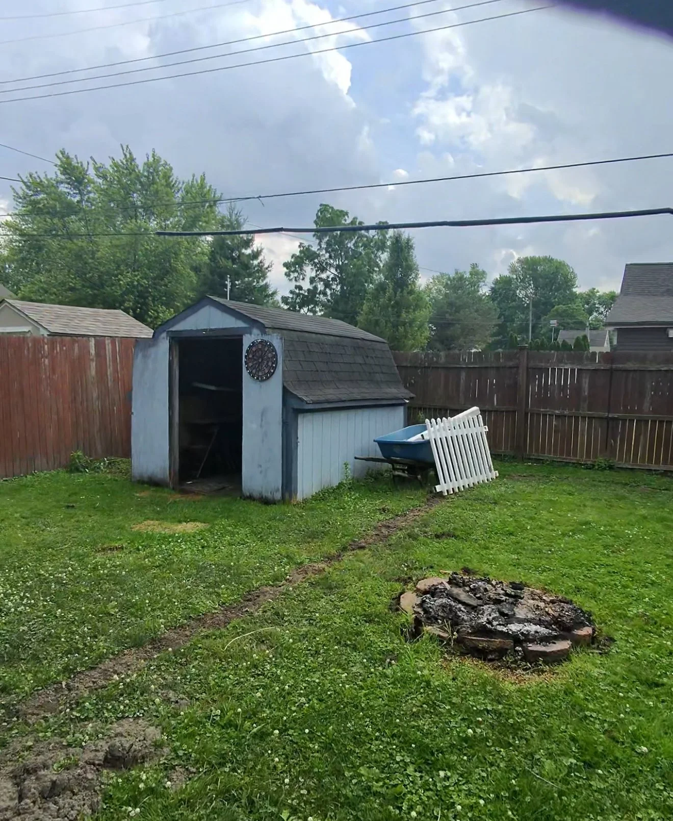 Backyard with a small shed, a wheelbarrow, a white picket fence piece, a fire pit, and a grassy area enclosed by a wooden fence, with trees and cloudy sky in the background.