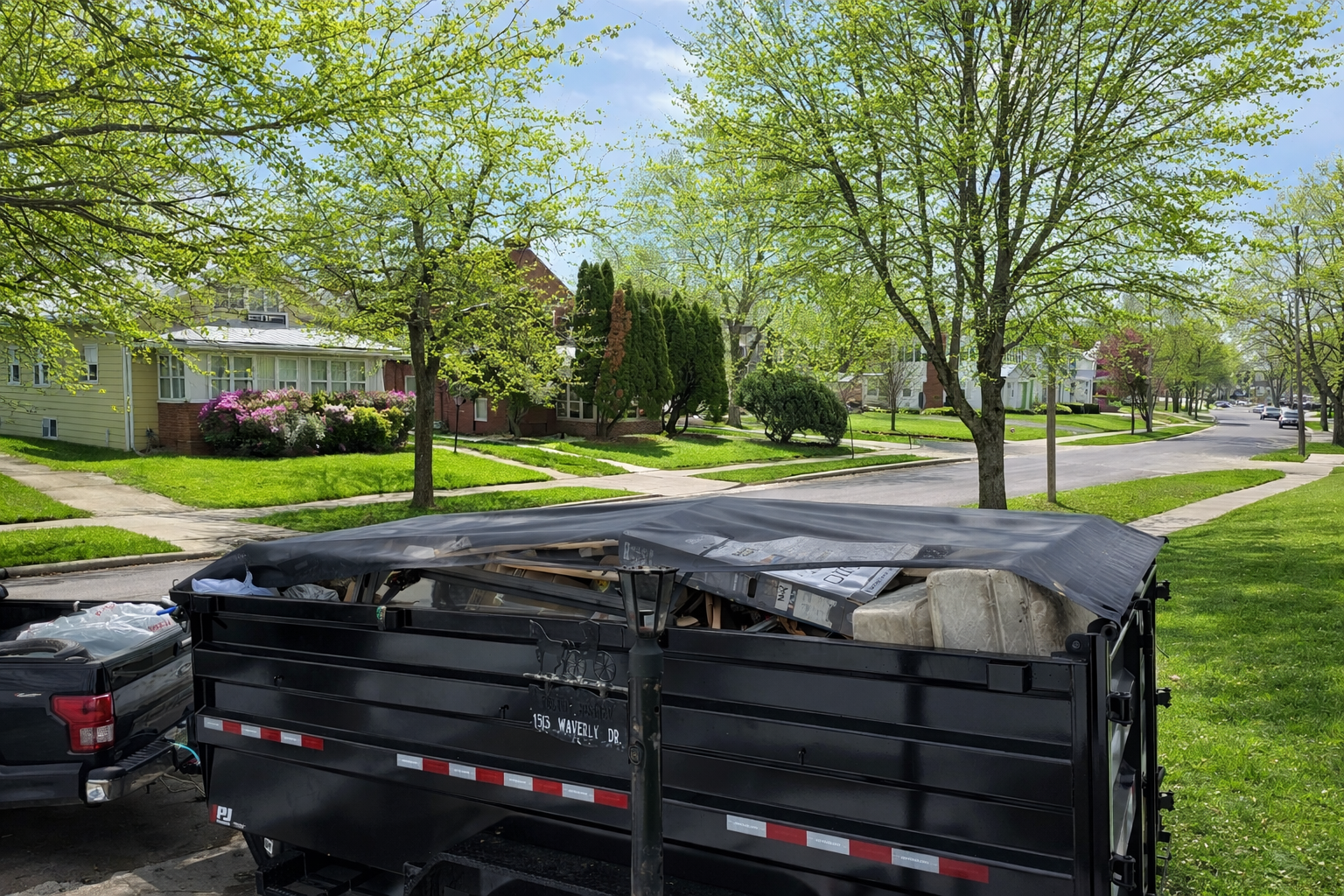 A residential neighborhood street lined with green trees and houses, with a moving truck filled with debris in the foreground.