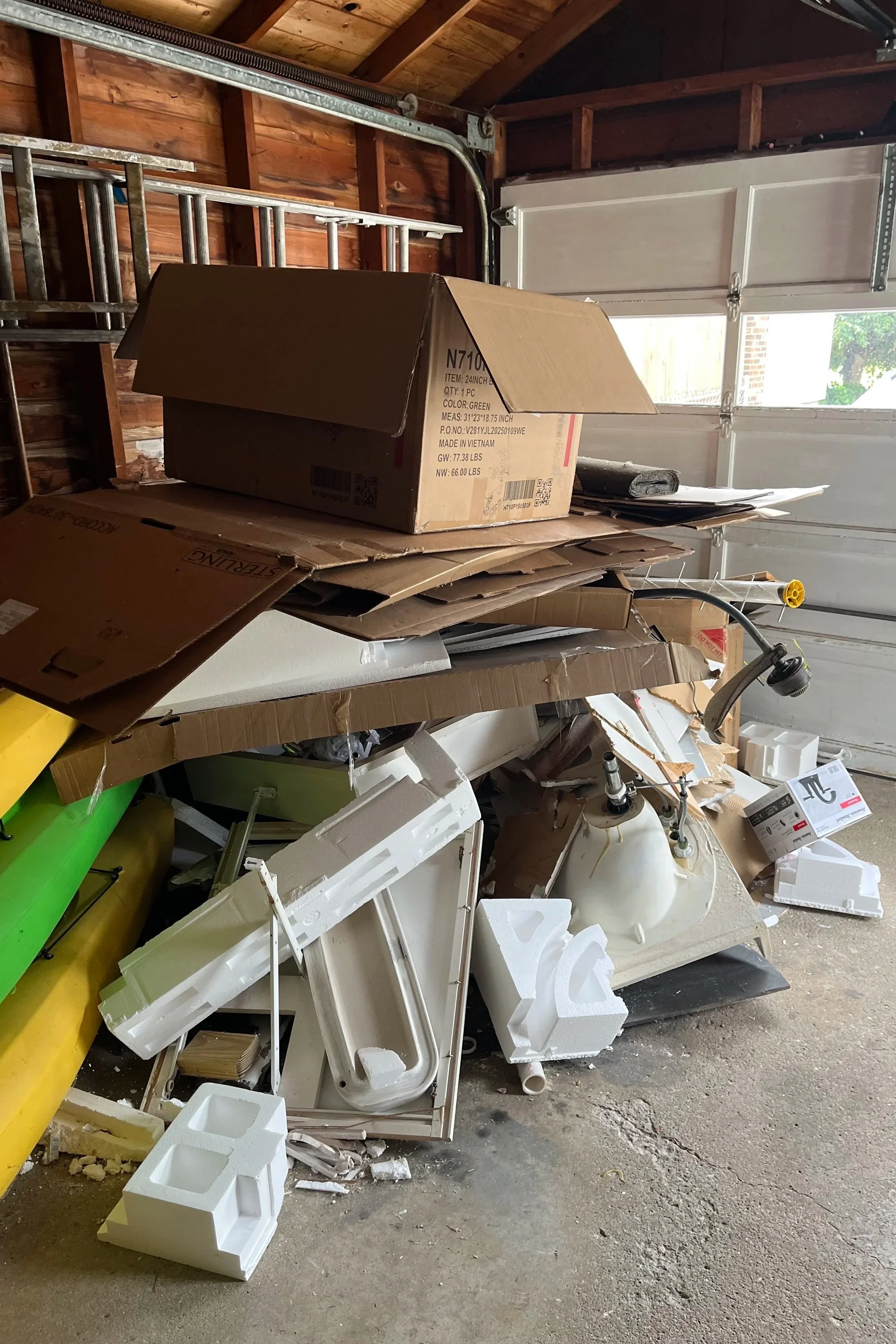 Piled-up items in a garage, including cardboard boxes, Styrofoam pieces, and a partially assembled light fixture or appliance, with a garage door and wooden walls in the background.