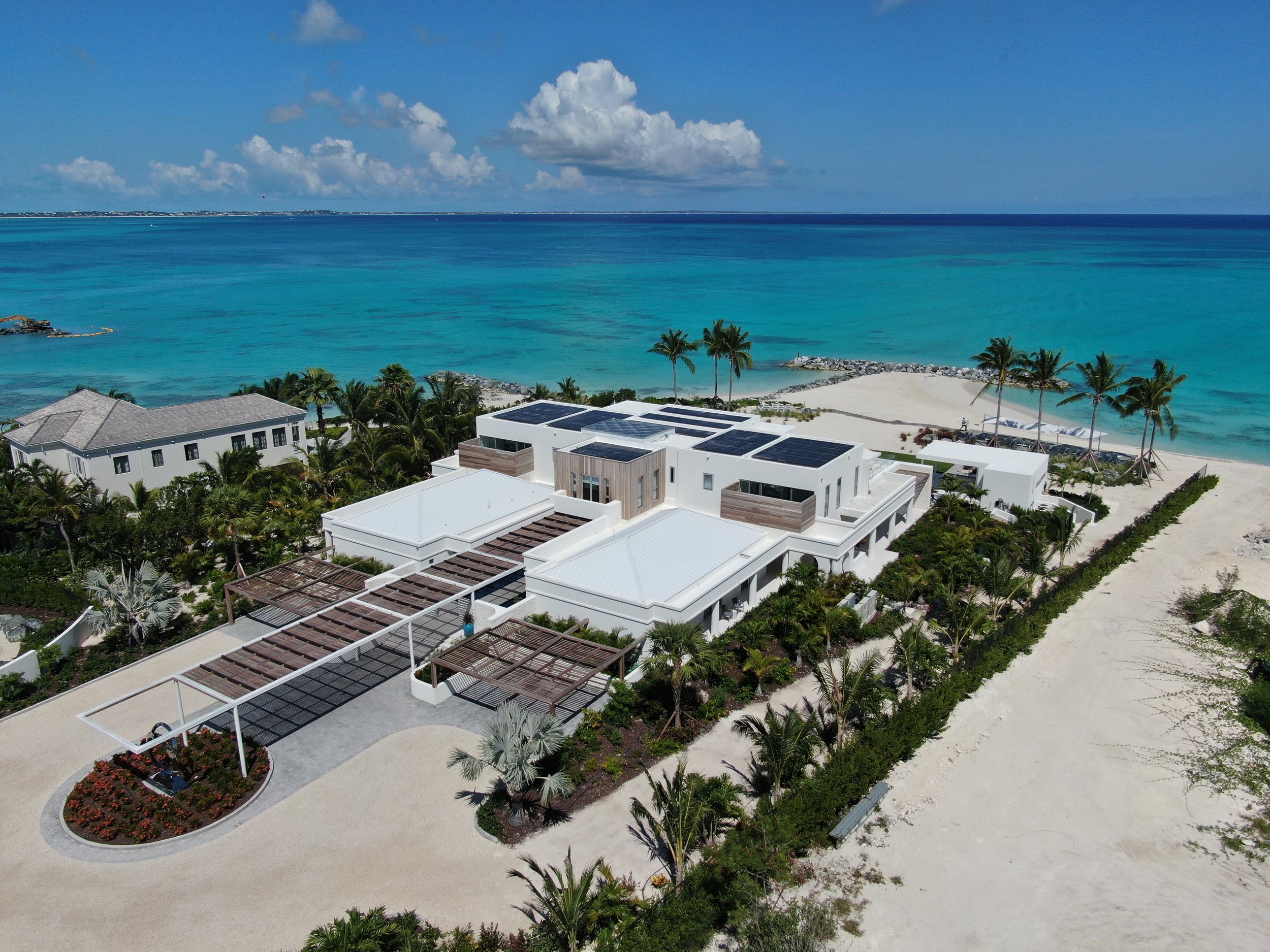 Aerial view of a coastal area with houses, a swimming pool, a tennis court, and a building complex near a sandy beach and turquoise ocean.