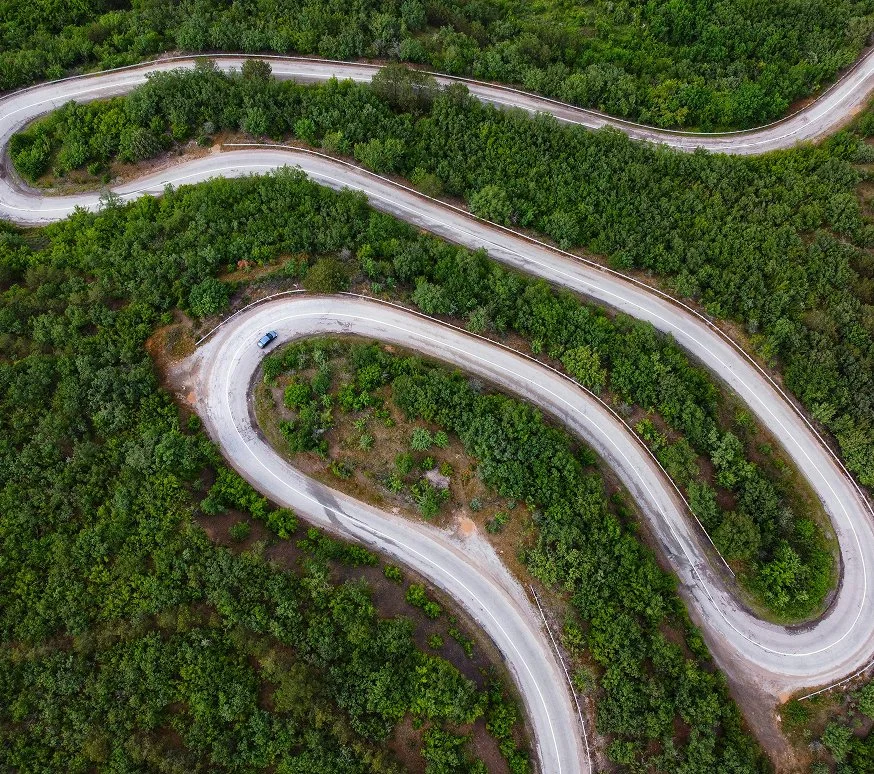 An aerial view of a winding road through a densely wooded area with a single blue car on the road.