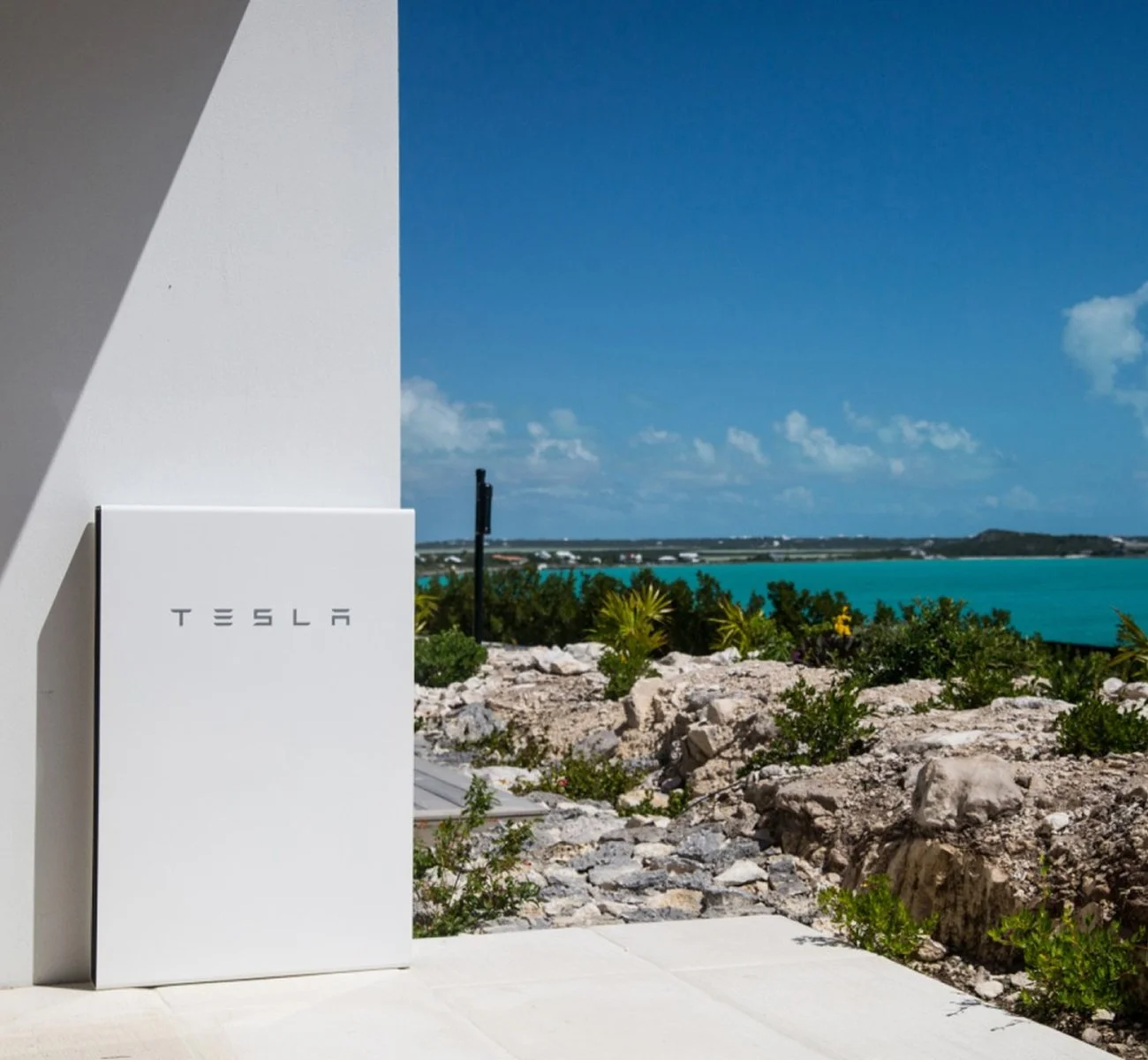 White box with the Tesla logo in black letters, situated outdoors on a concrete surface with a scenic view of a coastline, blue water, green plants, rocks, and a partly cloudy sky in the background.