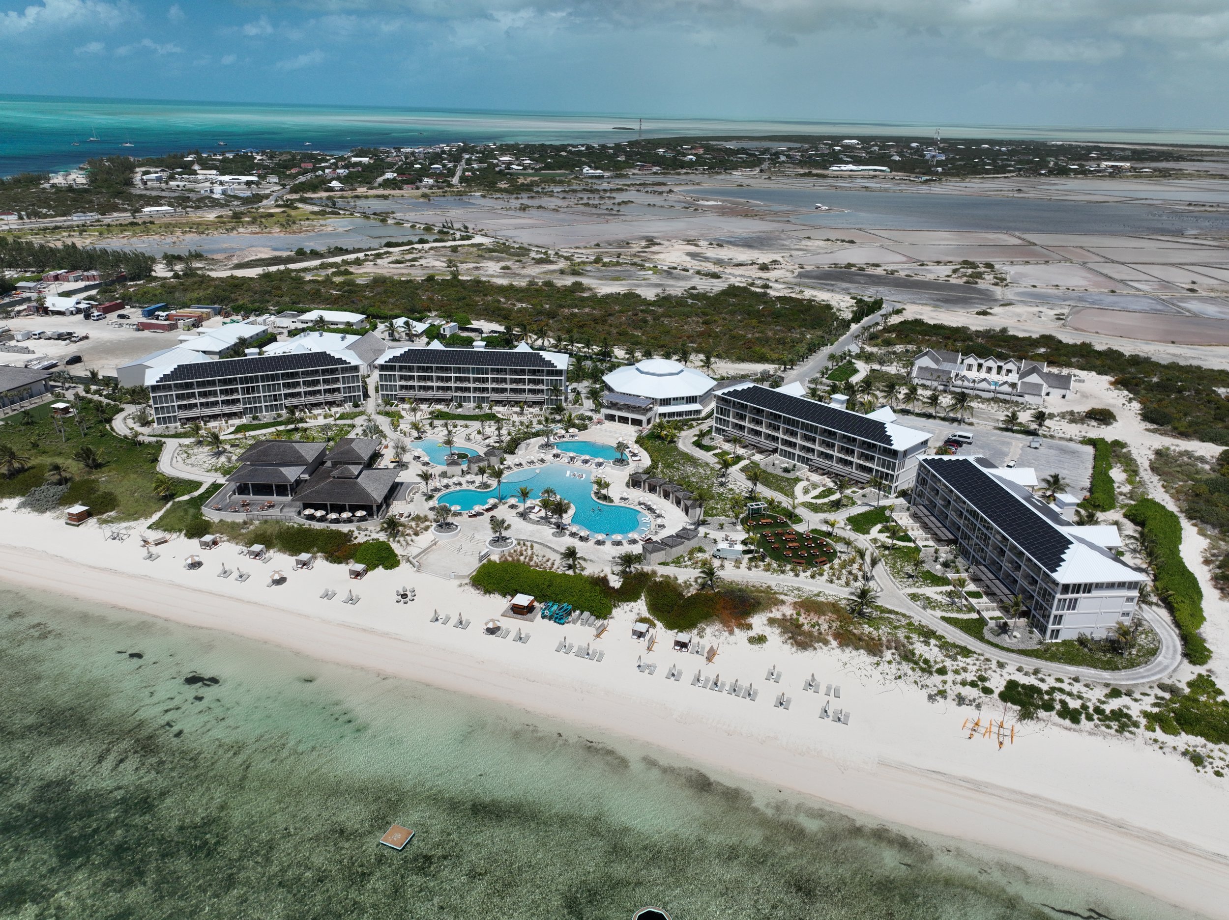 A large modern beachfront house with white walls and black solar panels on the roofs, surrounded by palm trees and lush greenery, located on a sandy beach with turquoise water and a breakwater in the distance.
