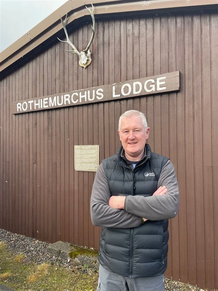 Lodge manager standing with arms crossed in front of a brown wooden building with a sign that says Rothiemurchus Lodge, and a mounted antlered animal skull above the sign. Article cover from Strathy Herald newspaper.