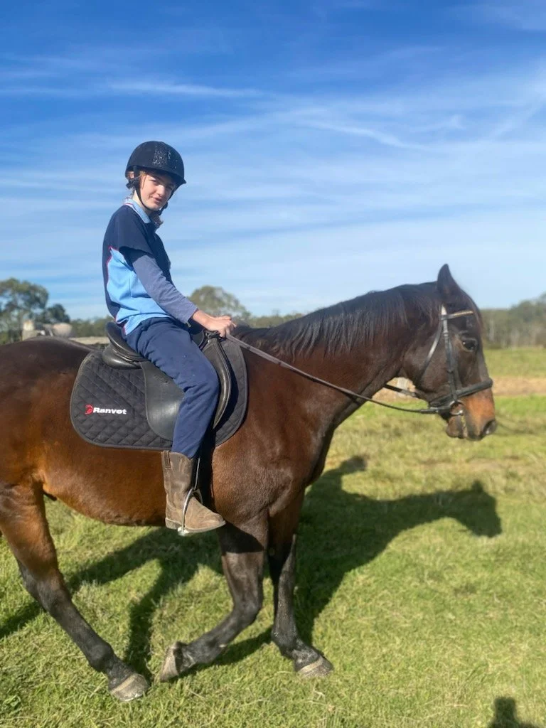 A young boy wearing a helmet and riding gear on a brown horse in an open field with a blue sky.