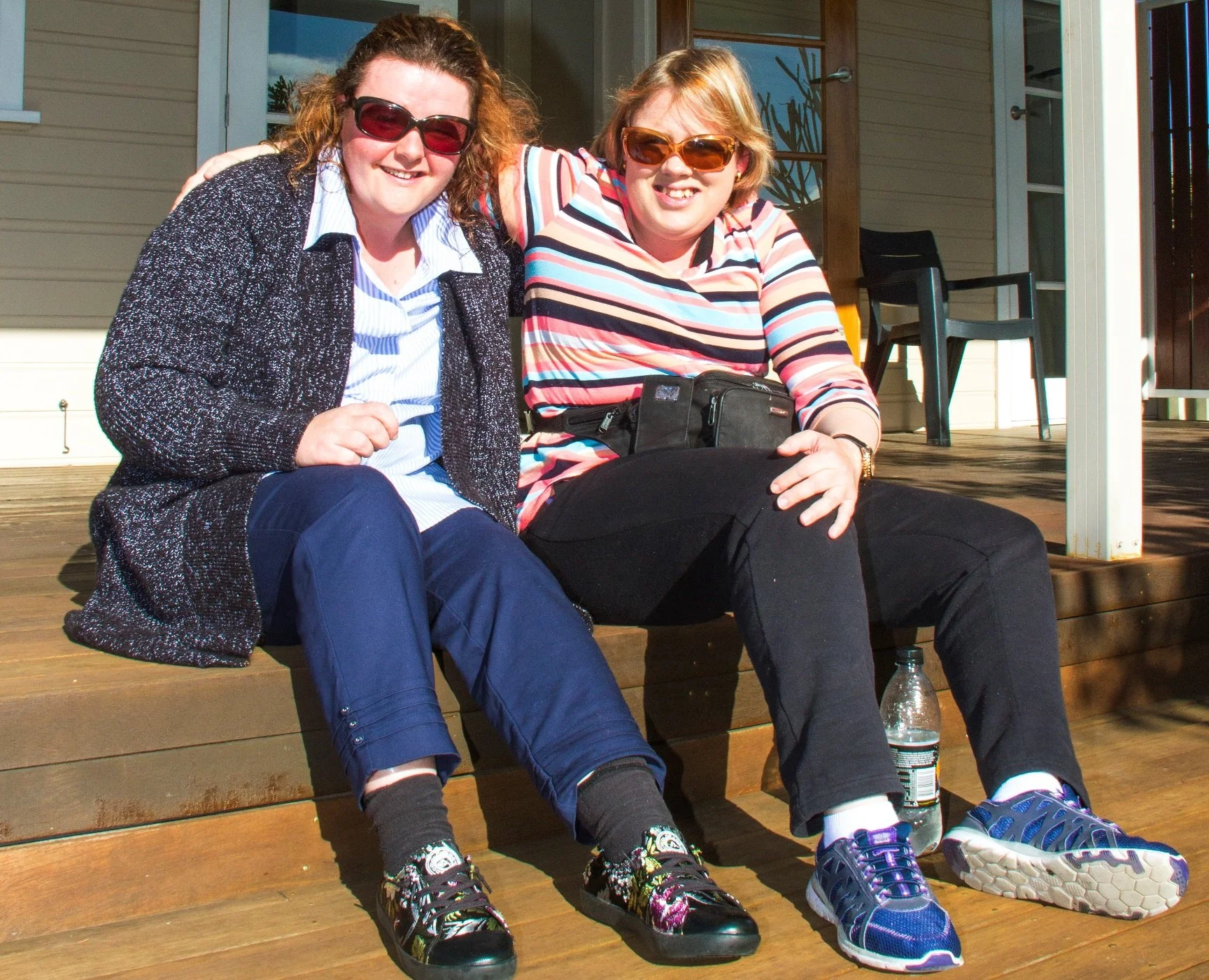 Two women sitting on a wooden porch, smiling and enjoying the sunny weather. One is wearing sunglasses, blue pants, and sneakers, while the other is wearing sunglasses, a striped long-sleeve shirt, black pants, and blue sneakers. 