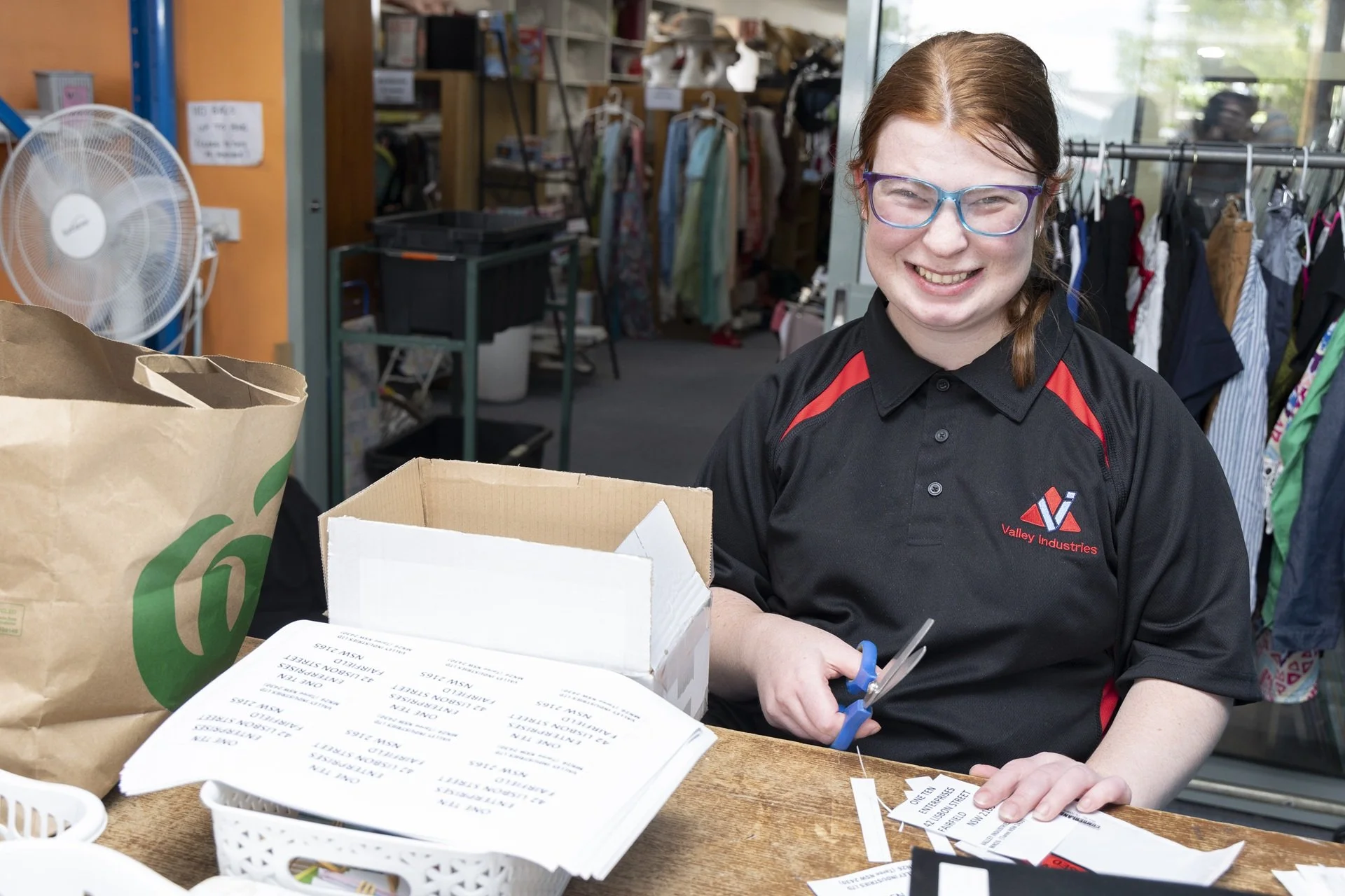 A team member with red hair, glasses, and a black shirt with red accents is smiling while cutting a paper tag with scissors at a counter. In the background, there are clothing racks, shelves, and a fan in our Valley Vintage shop.