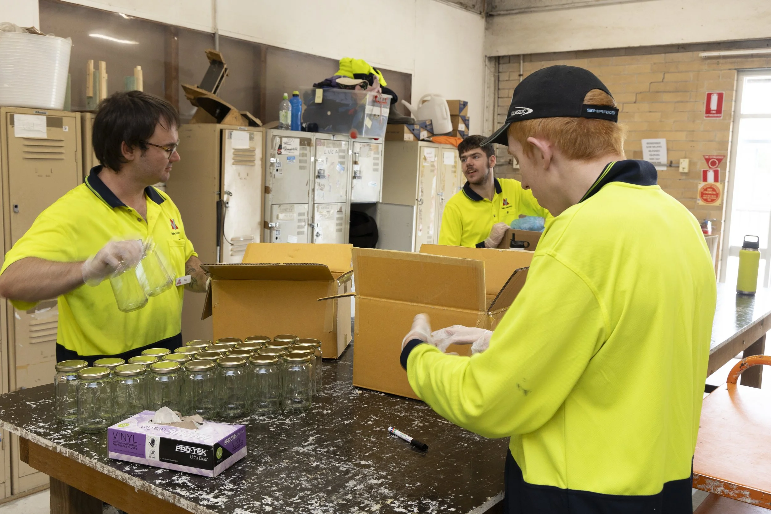 Four workers in high-visibility yellow shirts bagging jars in a workshop with lockers and supplies in the background.