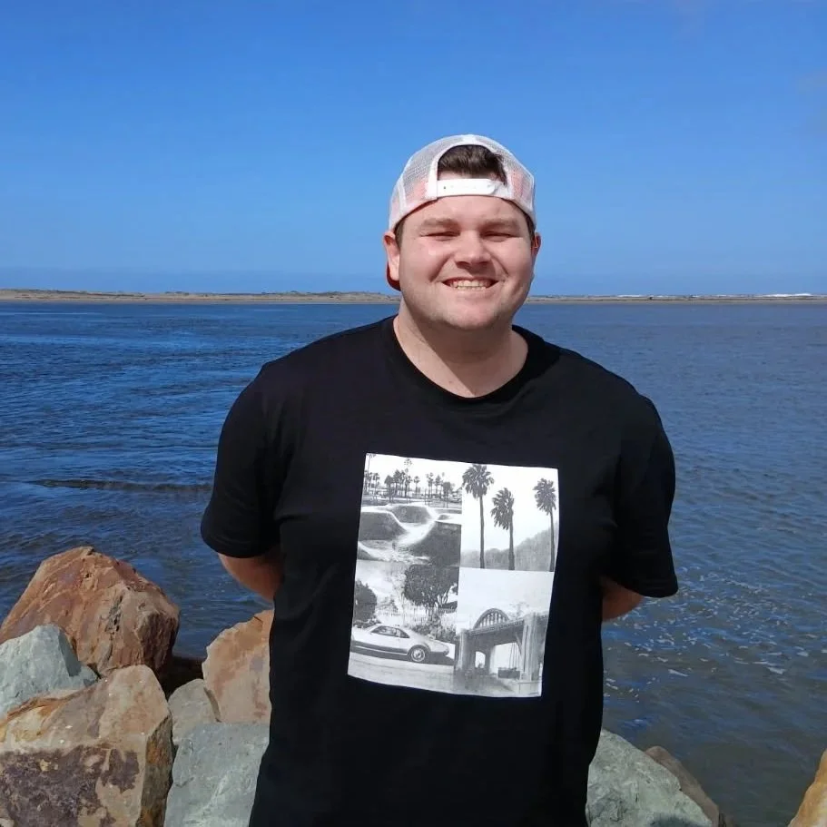 A young man smiling by the water with rocks, wearing a black t-shirt and a white baseball cap backward, on a sunny day.