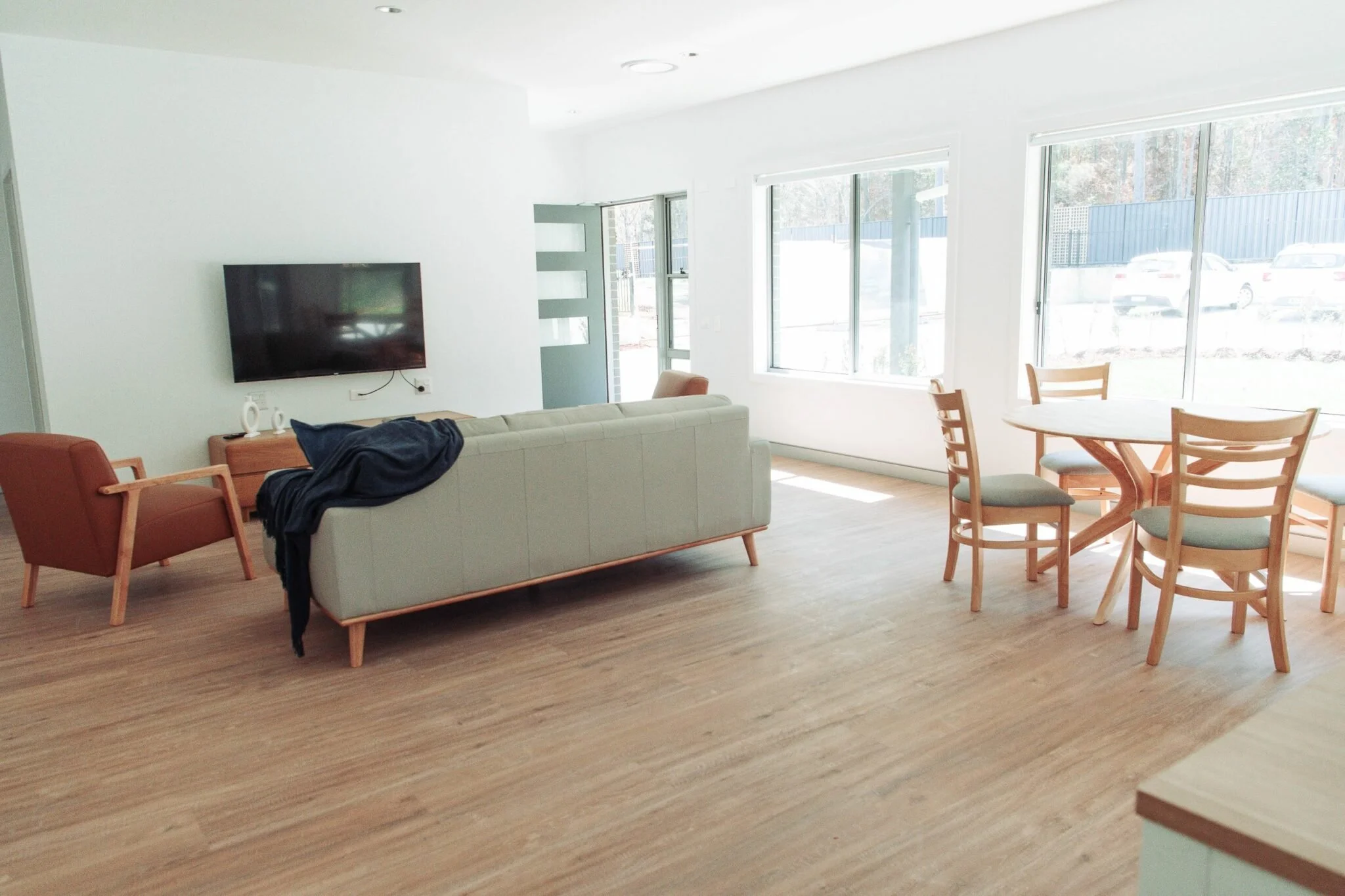 Bright living room with wooden flooring, a wall-mounted TV, a white sofa with a dark blue blanket, a coral-colored armchair, and a round wooden dining table with four chairs near large windows.