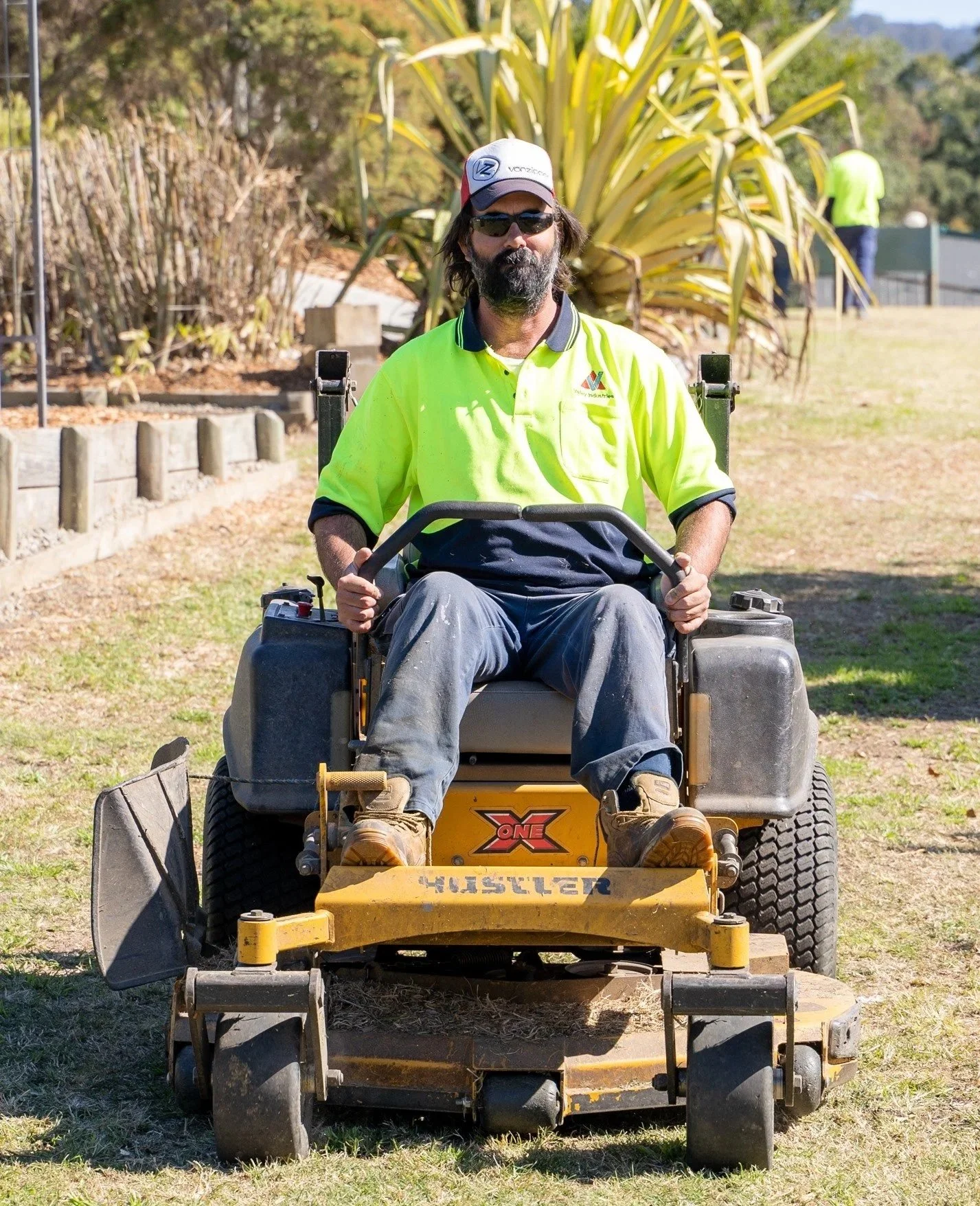 A man with a beard and sunglasses, wearing a high-visibility yellow and black work shirt, sitting on a lawn mower outdoors on a grassy area. There are plants and other people in the background.