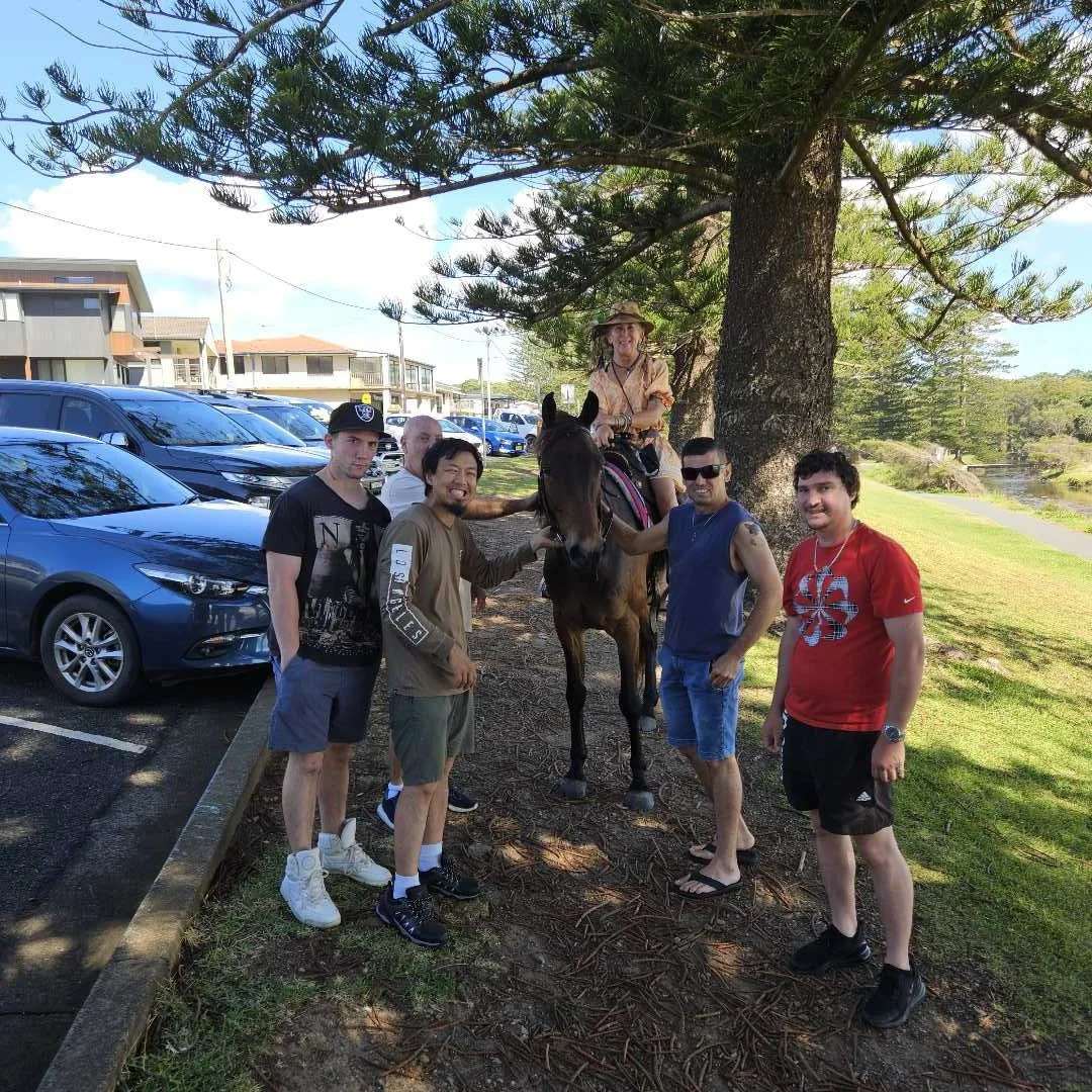 A group of people, including one on horseback, standing under a large tree in a park or recreational area, with parked cars in the background.