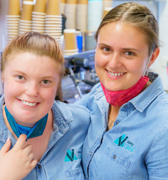 Two smiling female employees in light blue uniforms working at Valley TAFE Canteen, one with a face mask pulled down.