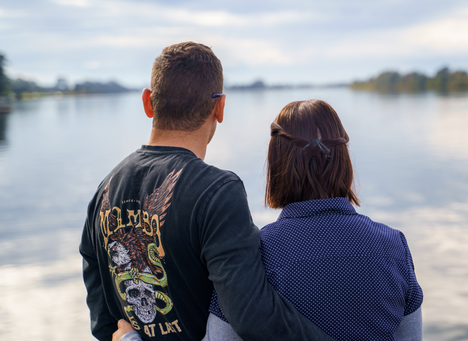 A man and woman sit together by a calm river, seen from behind, enjoying the peaceful scenery.
